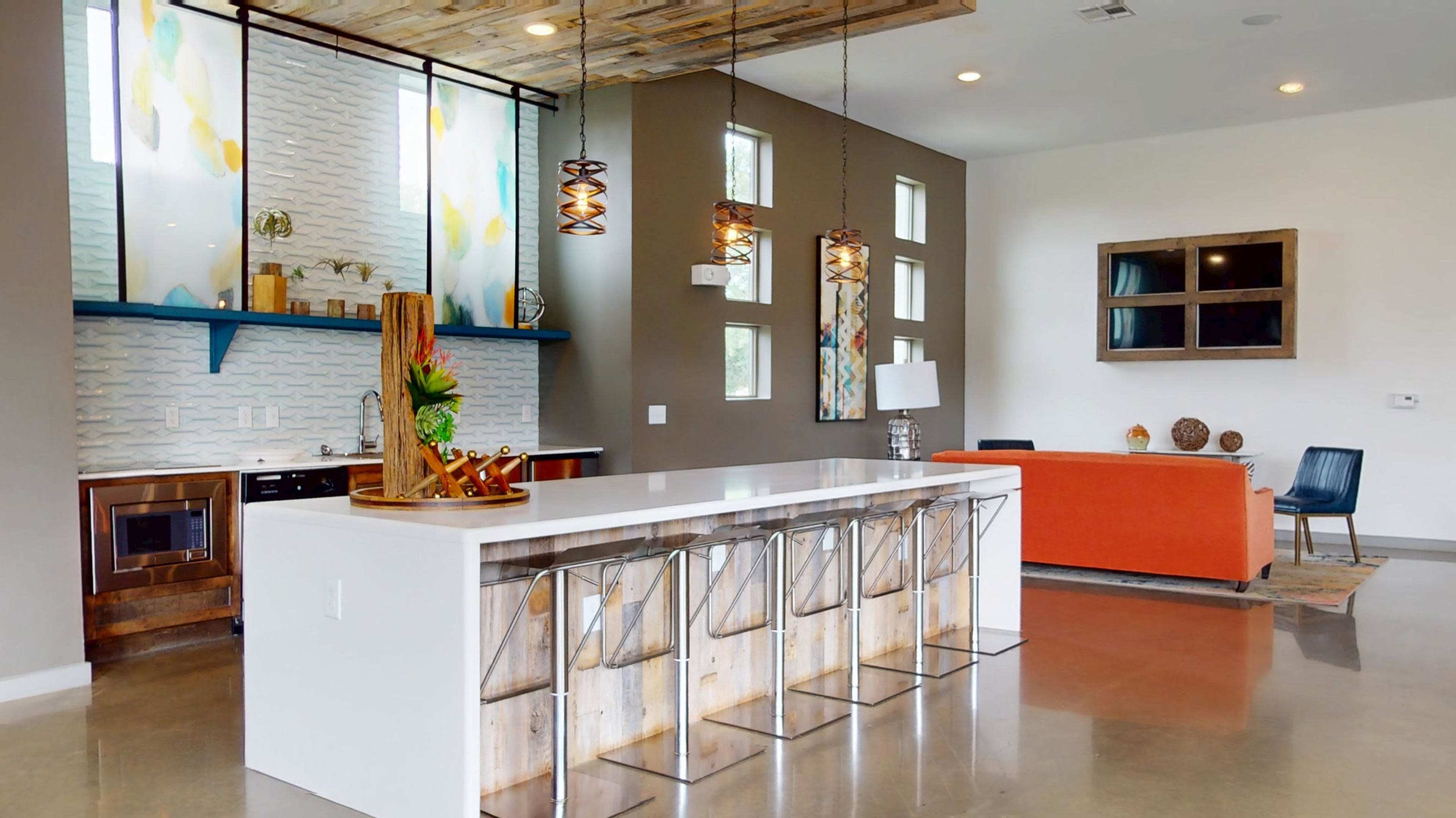 The image shows a modern kitchen and living area with a large white island, wooden bar stools, and a brightly colored orange sofa against a neutral wall.