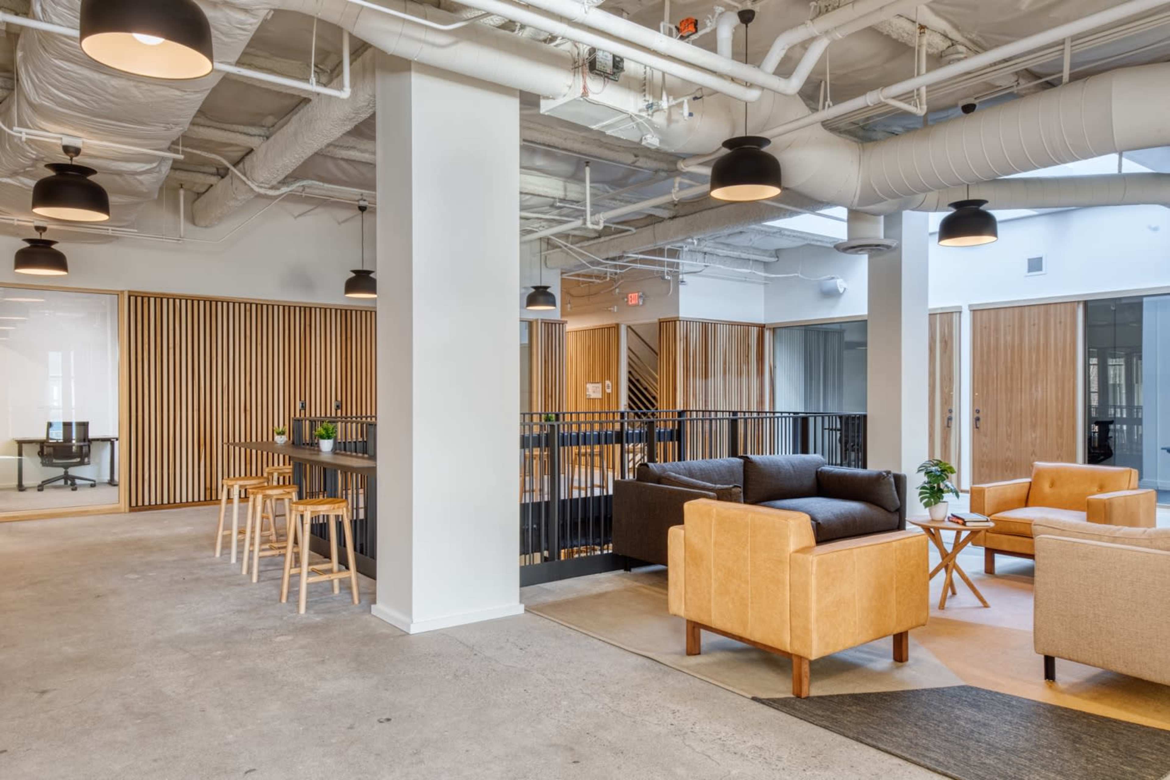 The image shows a modern open office space featuring wooden panels, seating areas with two armchairs, a couch, and a communal table with stools.