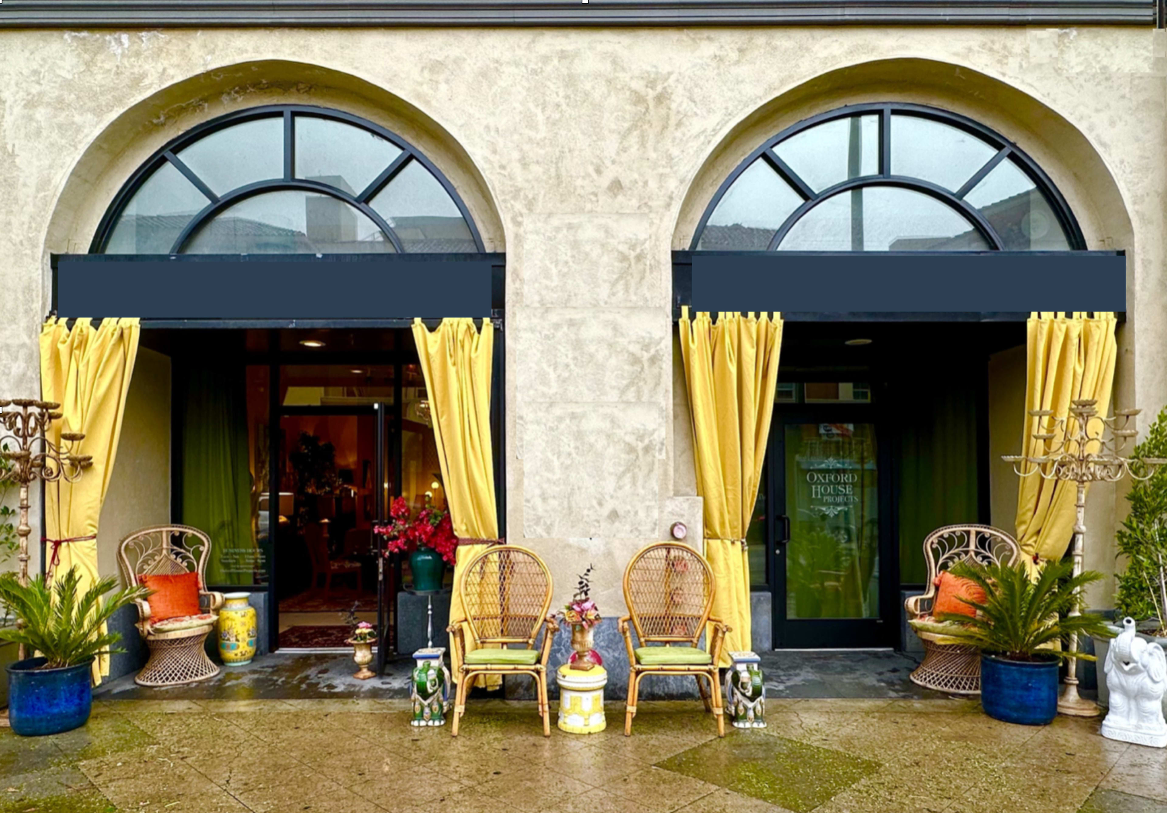 A building entrance with two arched doorways, each adorned with yellow curtains and flanked by potted plants and wicker chairs.