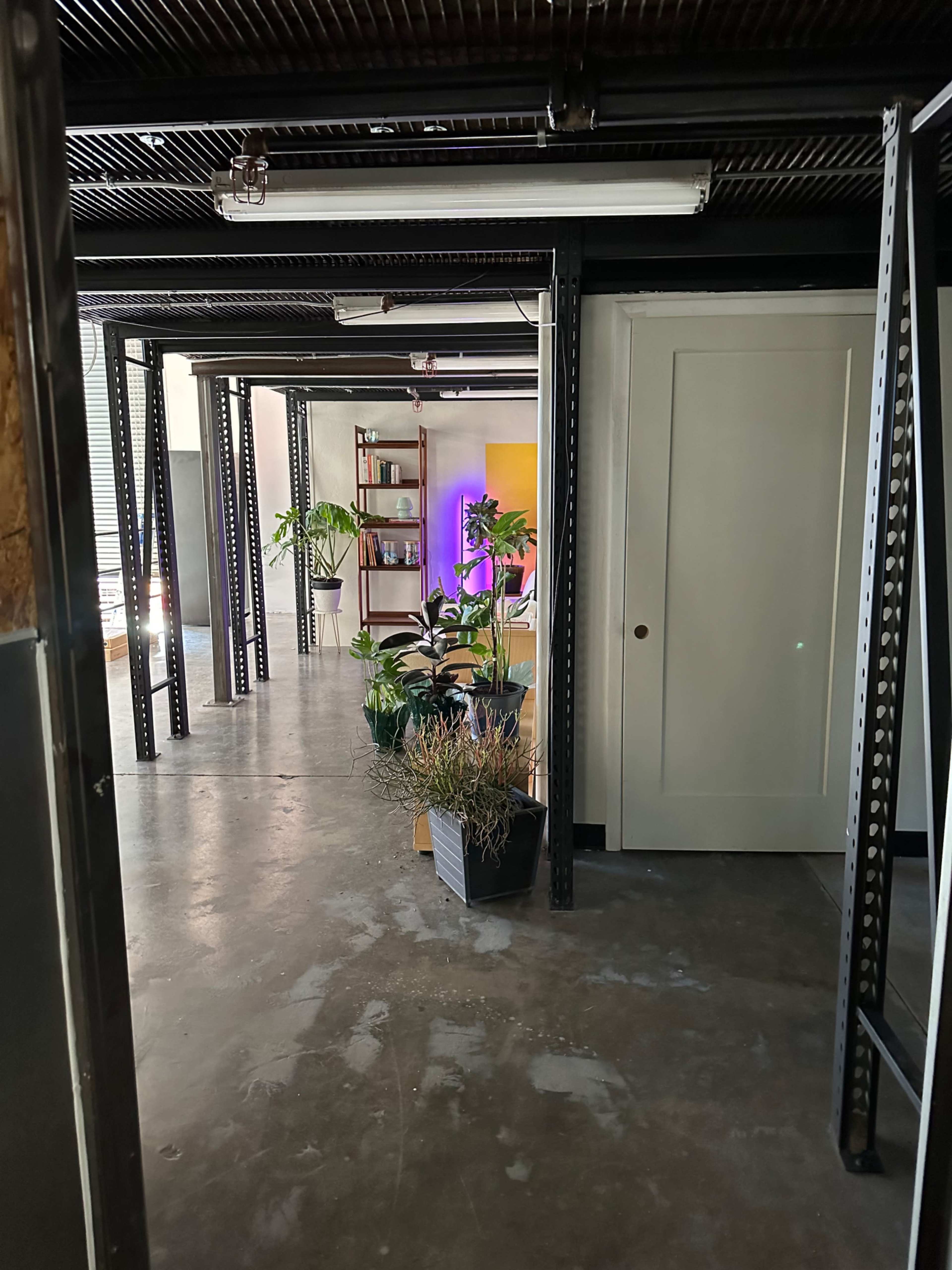 A hallway features industrial-style support beams leading towards a room filled with plants and shelves.