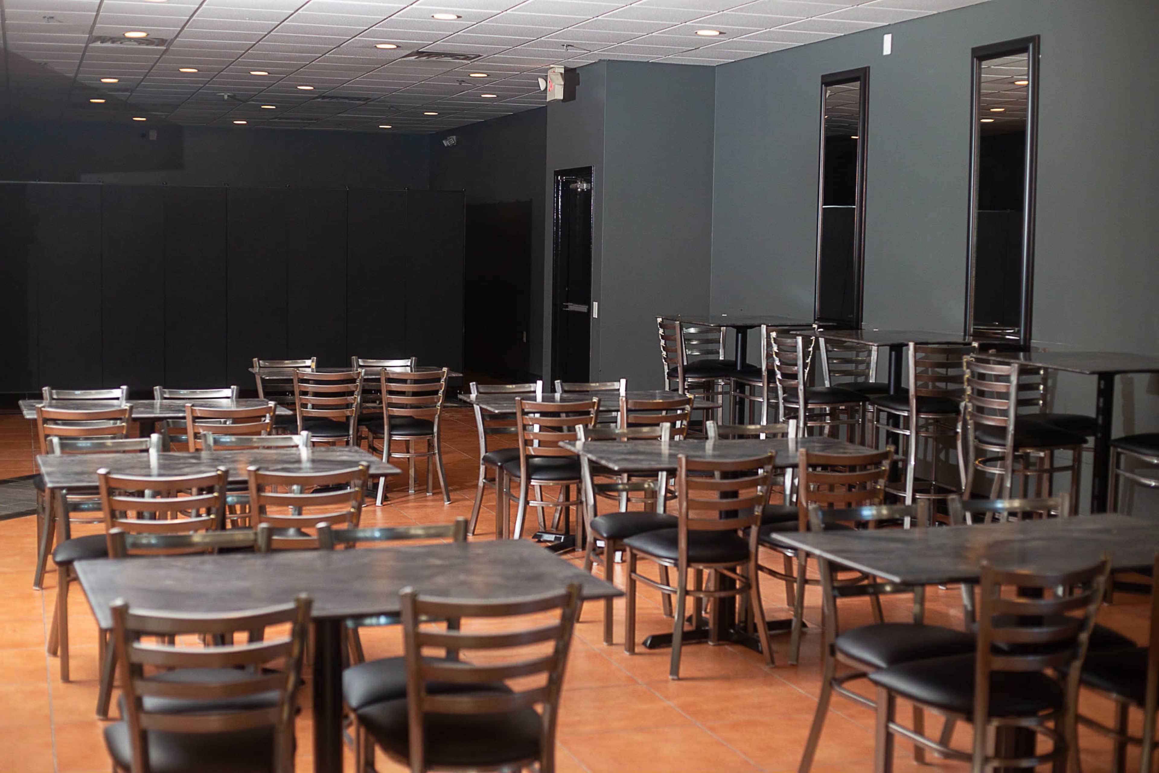 The image shows a spacious dining area with several empty tables and chairs arranged on a tile floor, featuring dark walls and minimal lighting.