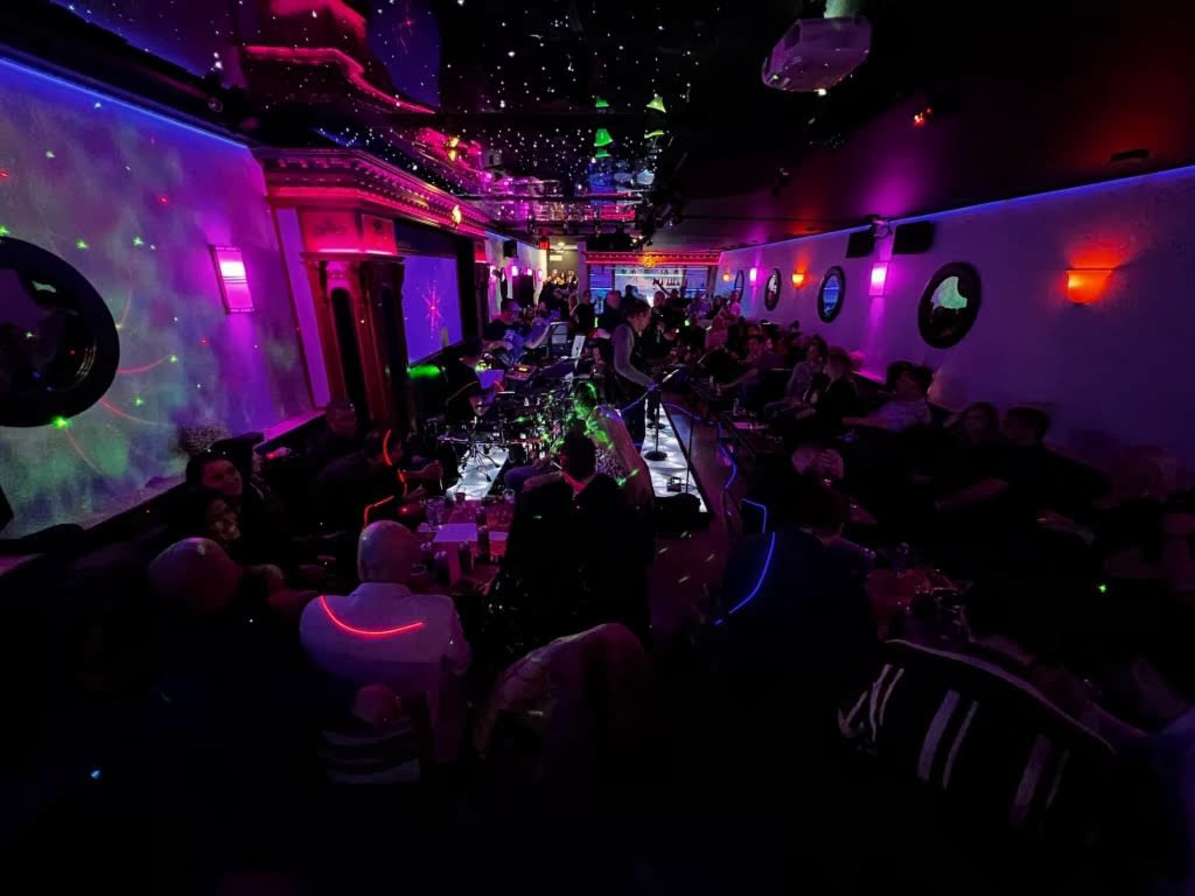 A lively bar scene is filled with patrons enjoying drinks under colorful lights and a starry ceiling.