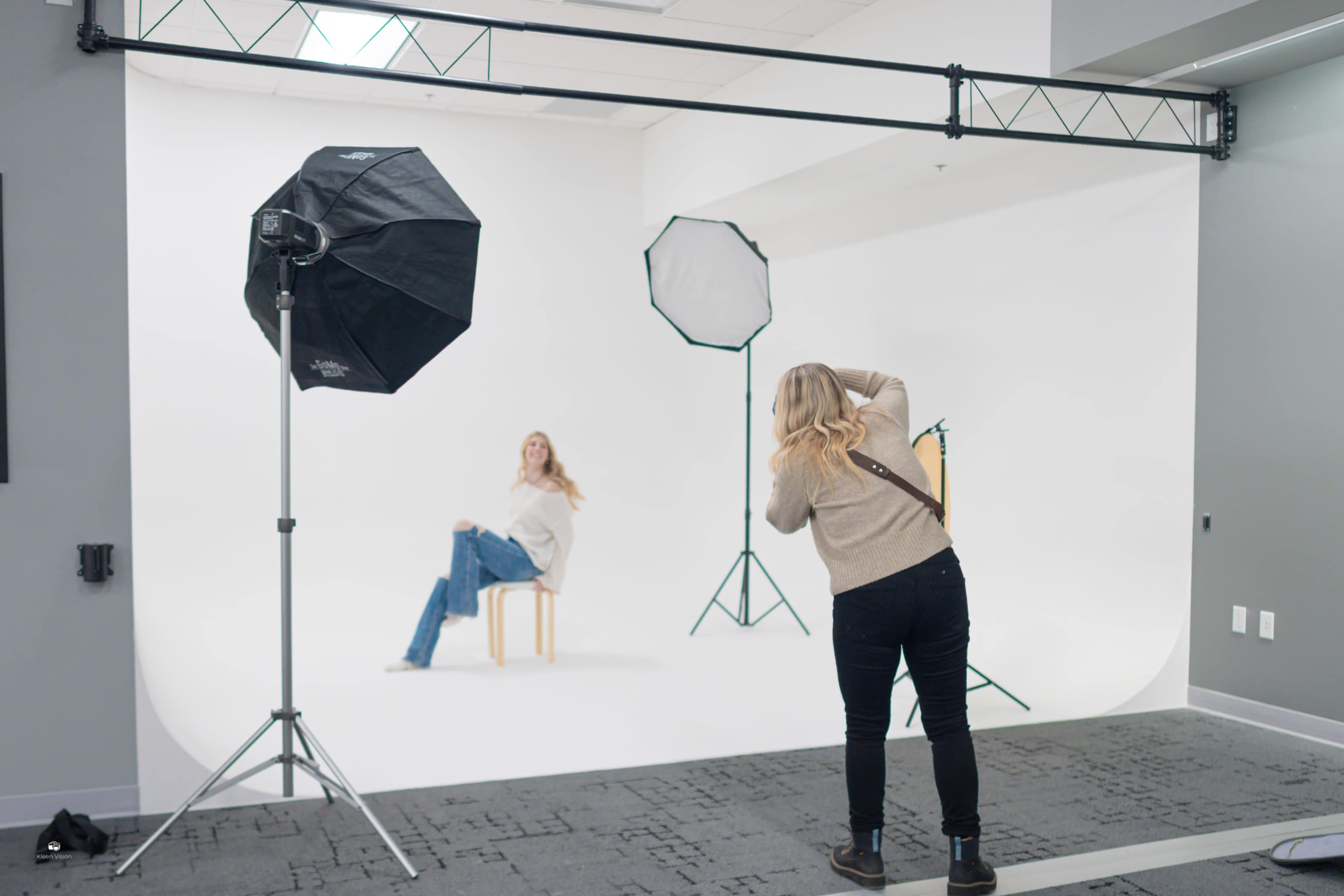 A photographer is taking pictures of a model seated on a stool in a well-lit studio with a simple white backdrop and lighting equipment.