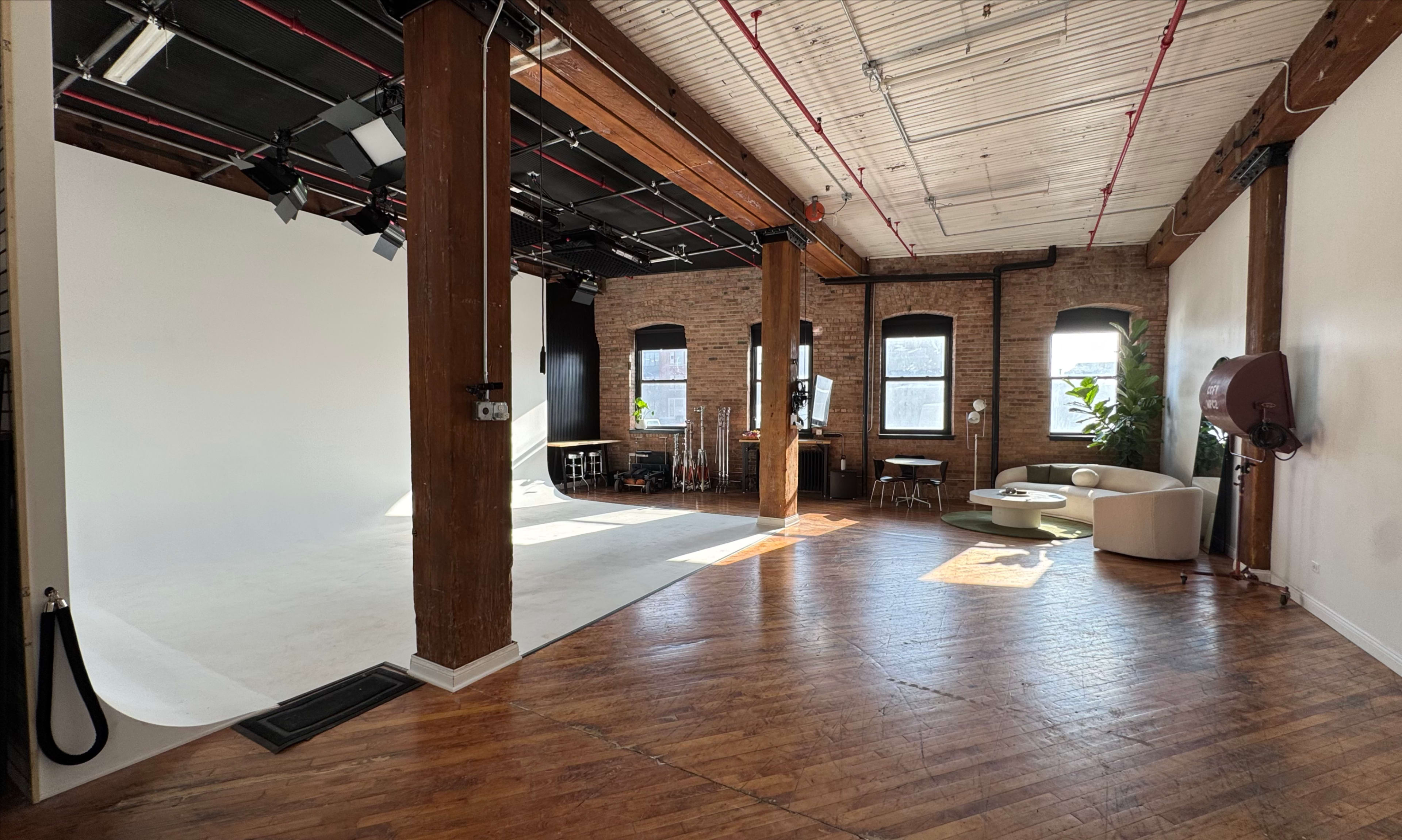 The image shows a spacious photography studio with wooden floors, exposed brick walls, and large windows, featuring a white backdrop and various pieces of furniture.