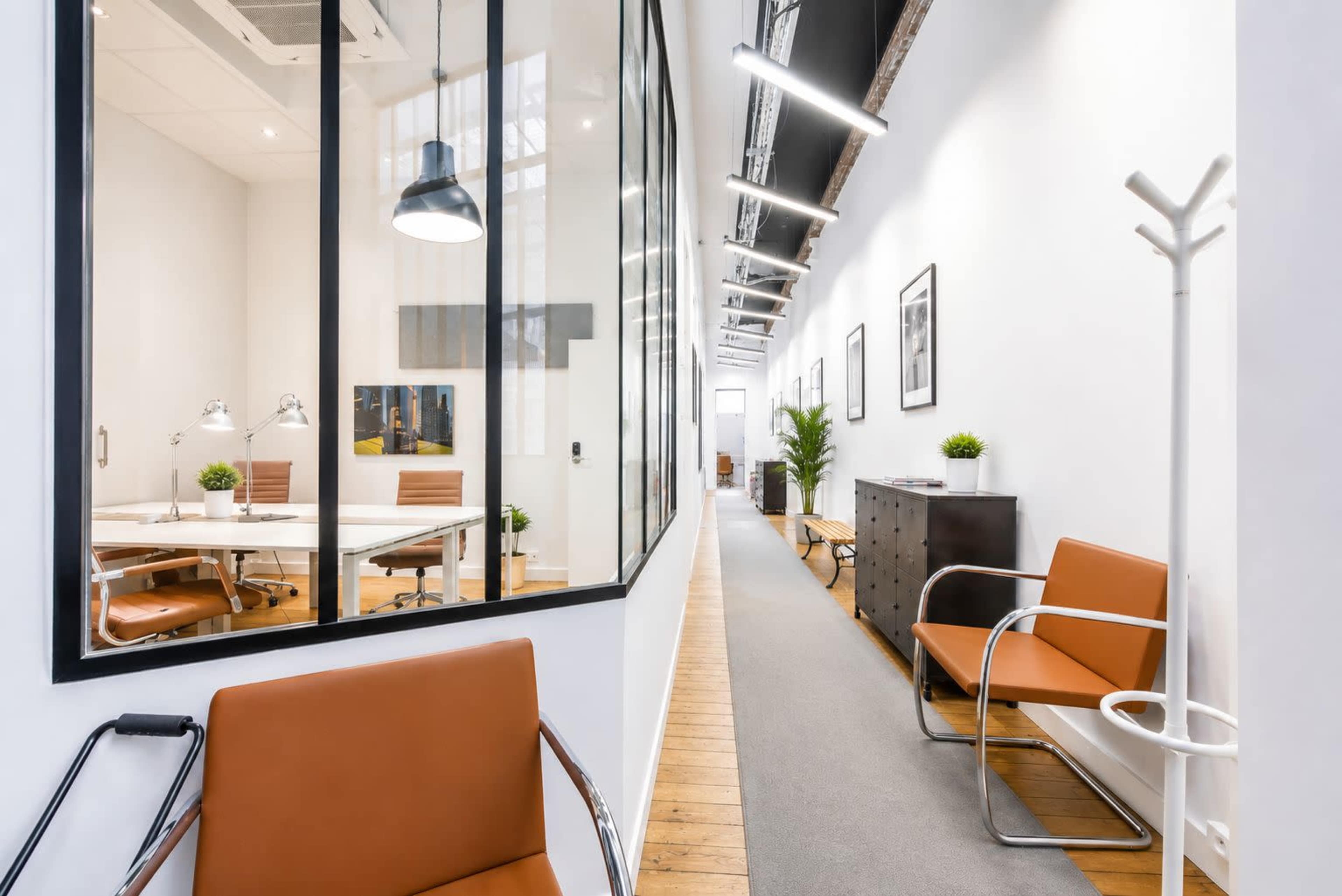 The image shows a modern office corridor with a glass-walled conference room on one side and a well-furnished seating area on the other.