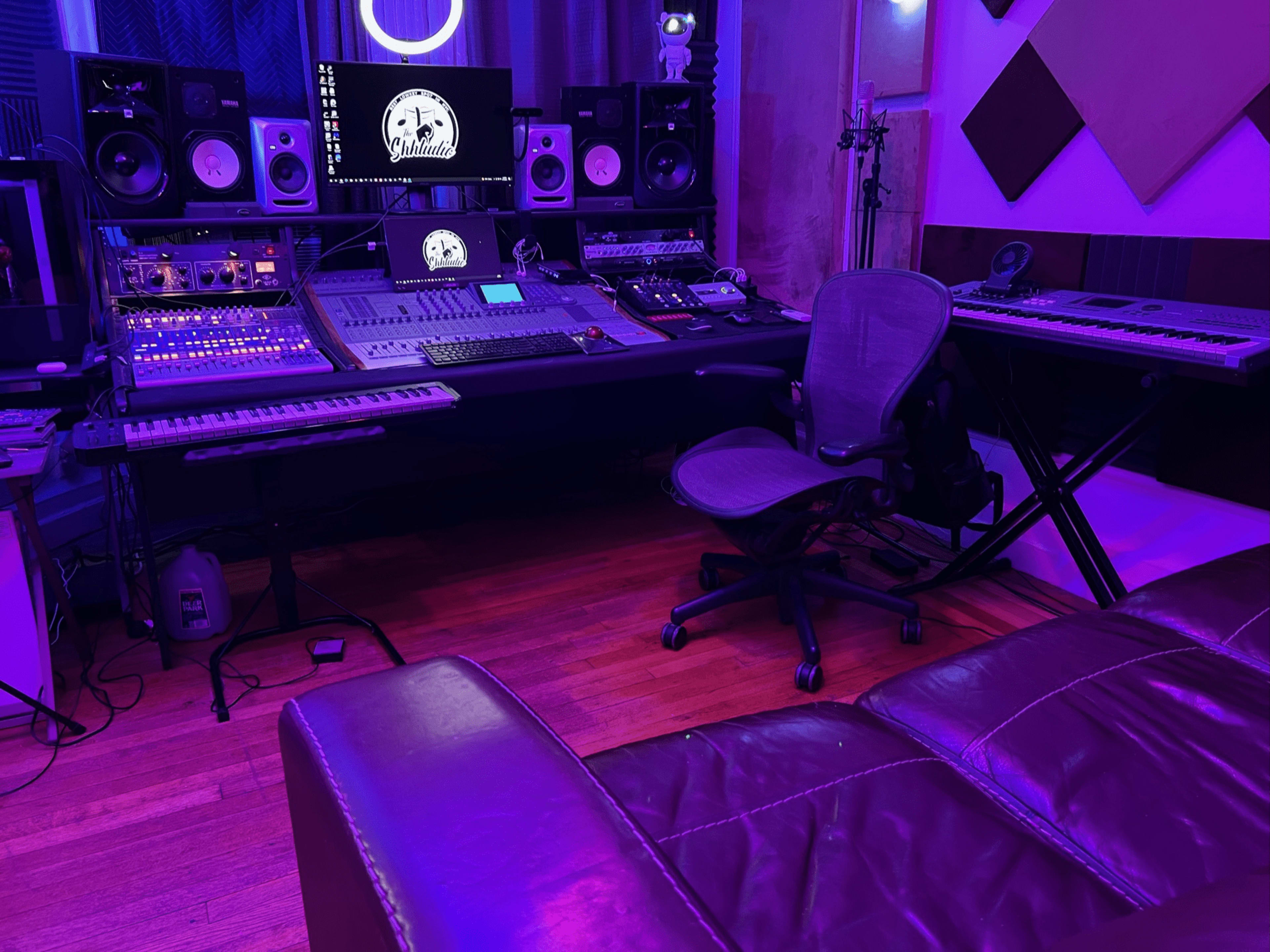A modern music studio with a mixing console, multiple speakers, keyboards, and a leather couch under purple lighting.