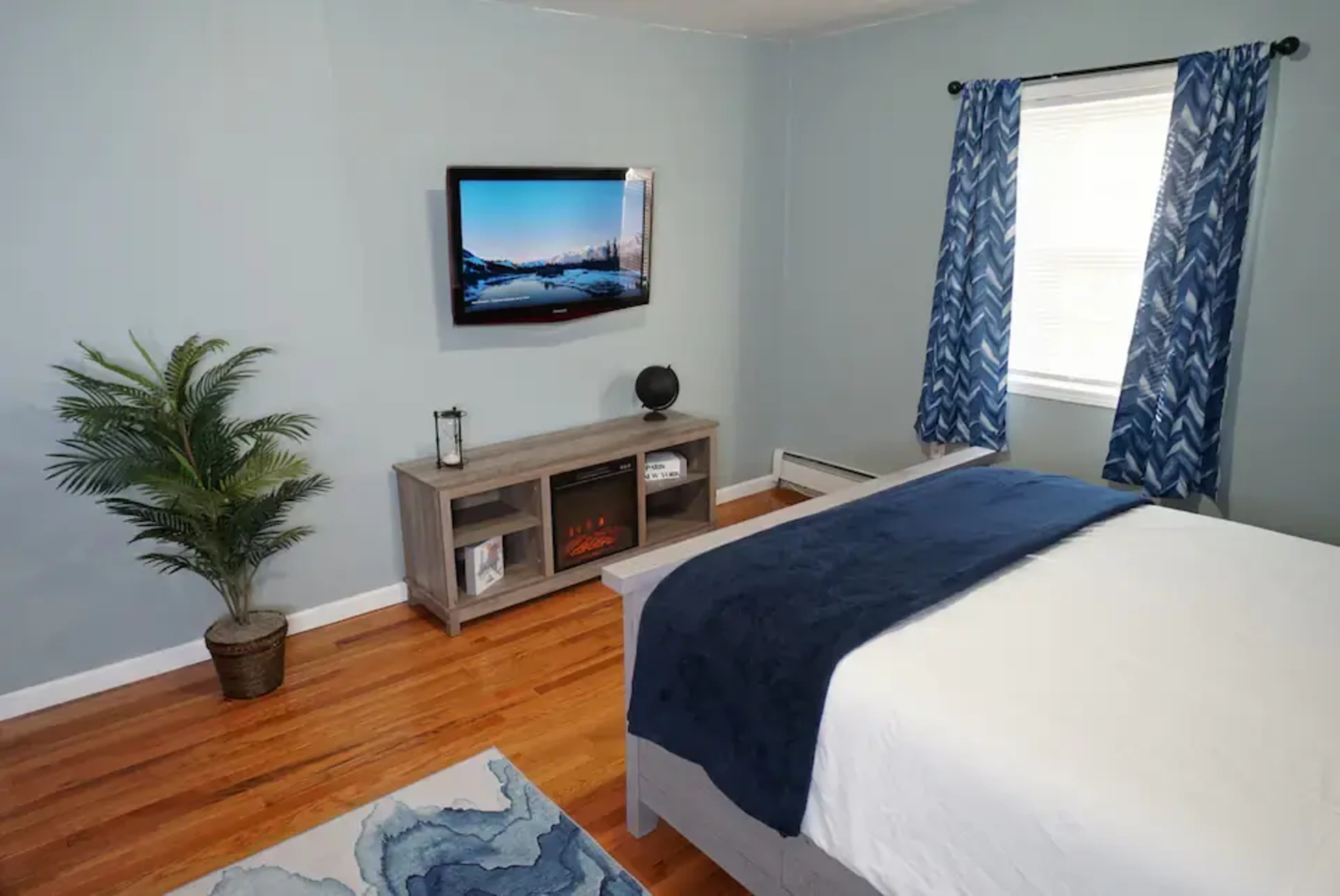 A bedroom with blue walls, hardwood flooring, a television mounted on the wall, a bed with a dark blue blanket, and a potted plant in the corner.
