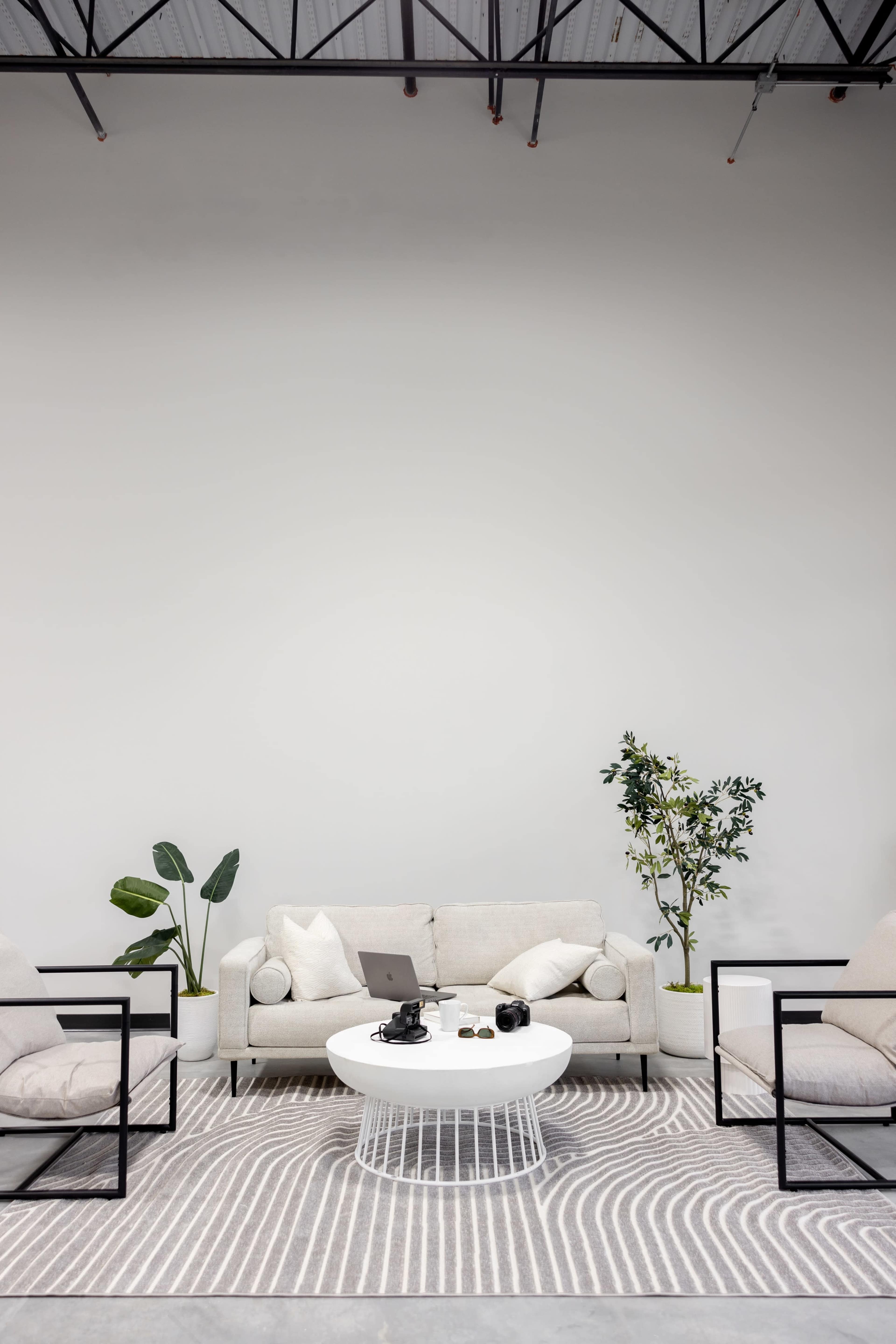 A modern lounge area features a light-colored couch, two black chairs, and a low coffee table, surrounded by indoor plants against a plain wall.