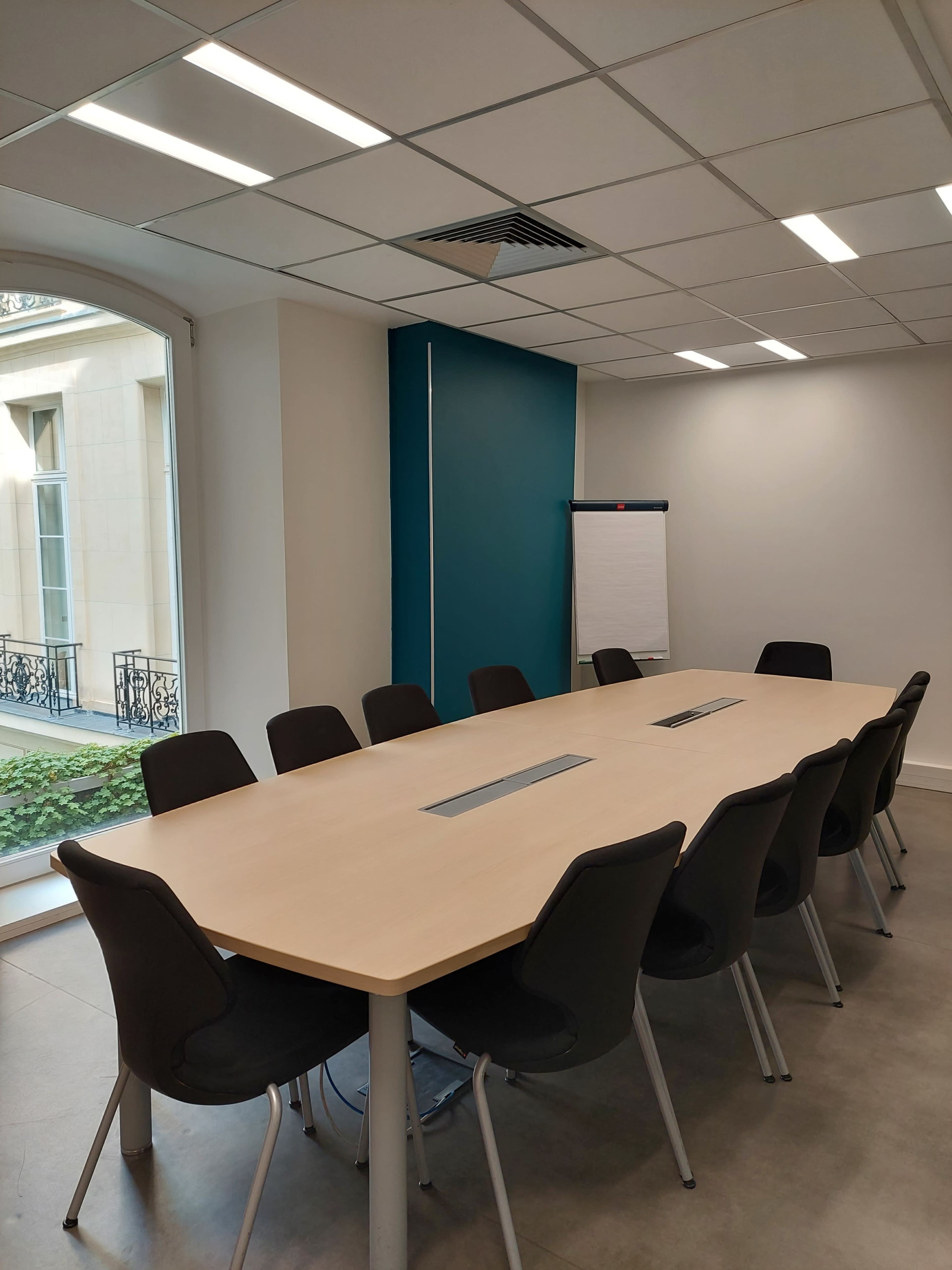 A large, modern conference room features a long table surrounded by chairs, with a flip chart on one side and windows providing natural light.