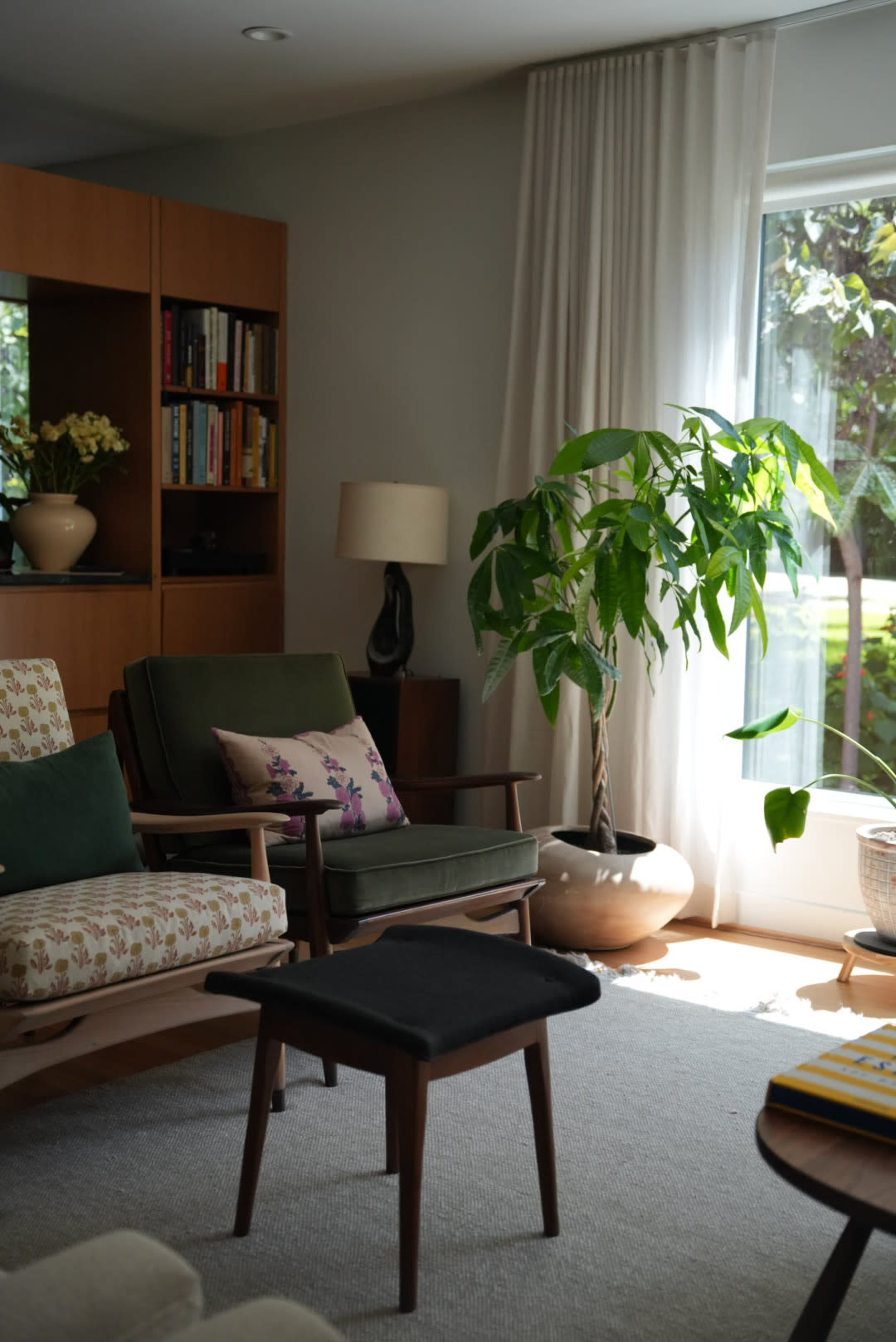 The living room features a pair of armchairs, a small stool, a bookcase, and a large window with a potted plant nearby.