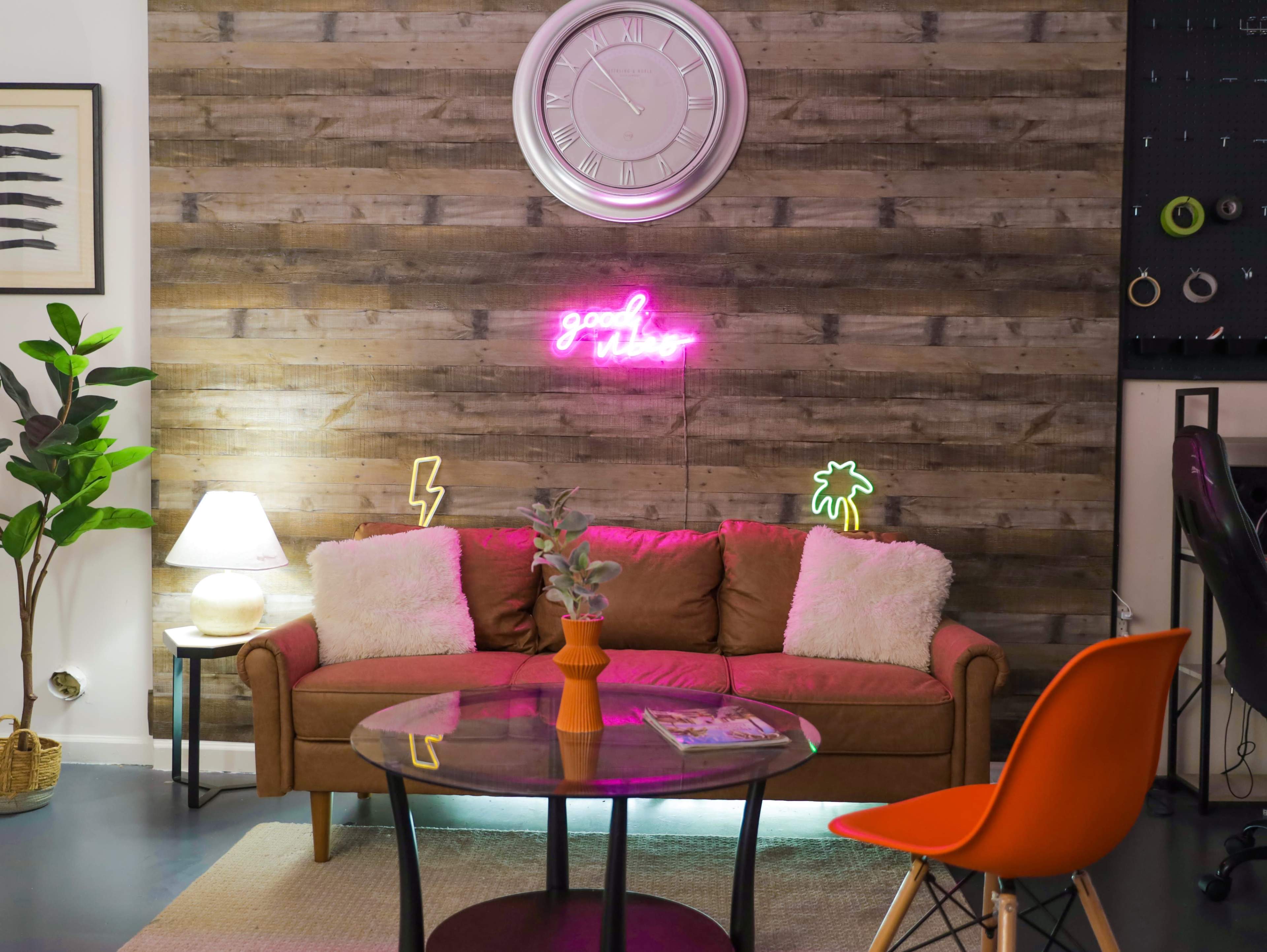 A cozy living room features a brown sofa with plush cushions, a round glass coffee table, and a wall adorned with a neon sign and decorative plants.