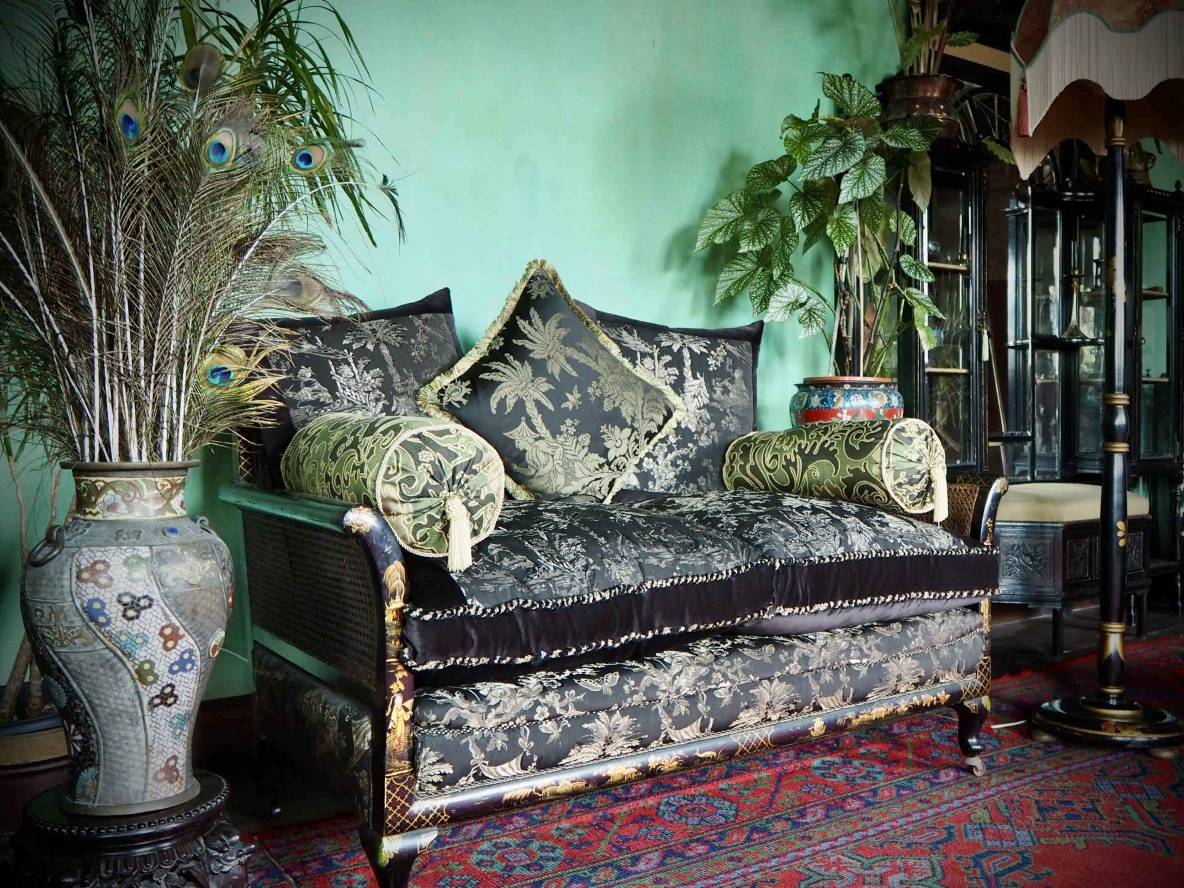 The image features a luxurious black and gold upholstered settee with decorative cushions, positioned against a green wall and surrounded by plants and ornate furnishings.