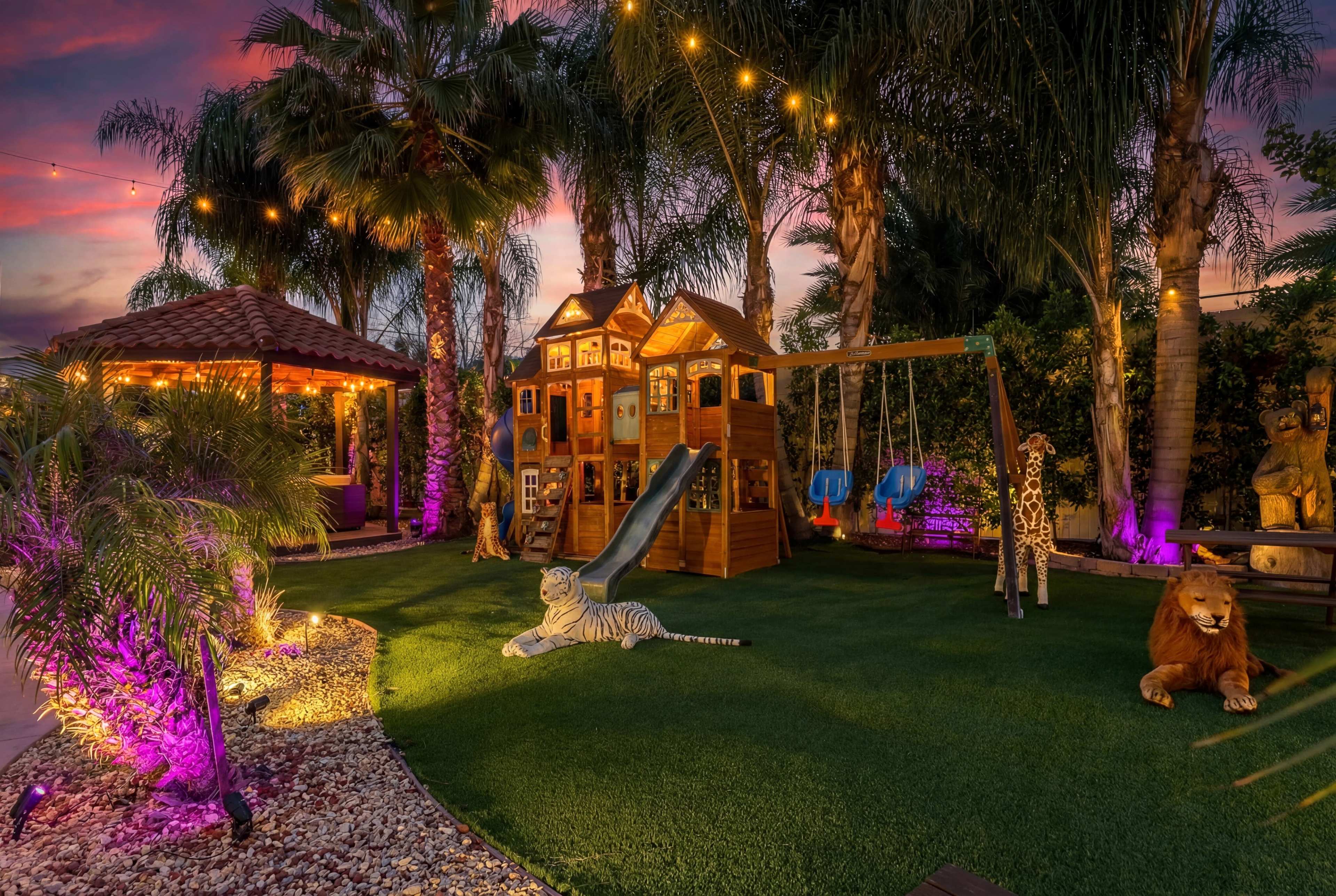 The image shows a backyard play area featuring a wooden playhouse, swings, a slide, and decorative animal statues, all under string lights and surrounded by palm trees.