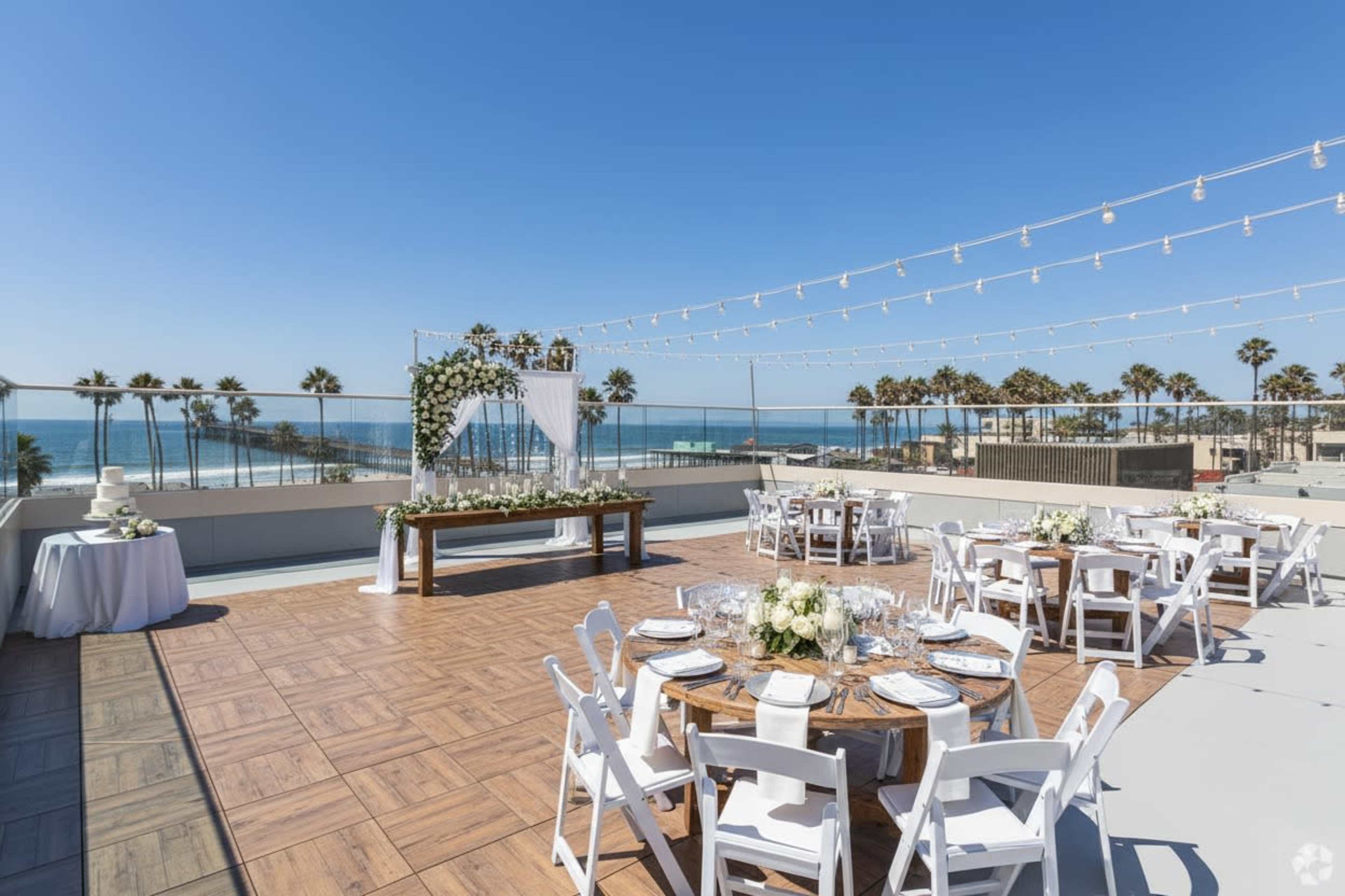 The image shows a decorated rooftop venue overlooking the ocean, featuring tables set for a gathering with white chairs and a floral backdrop.