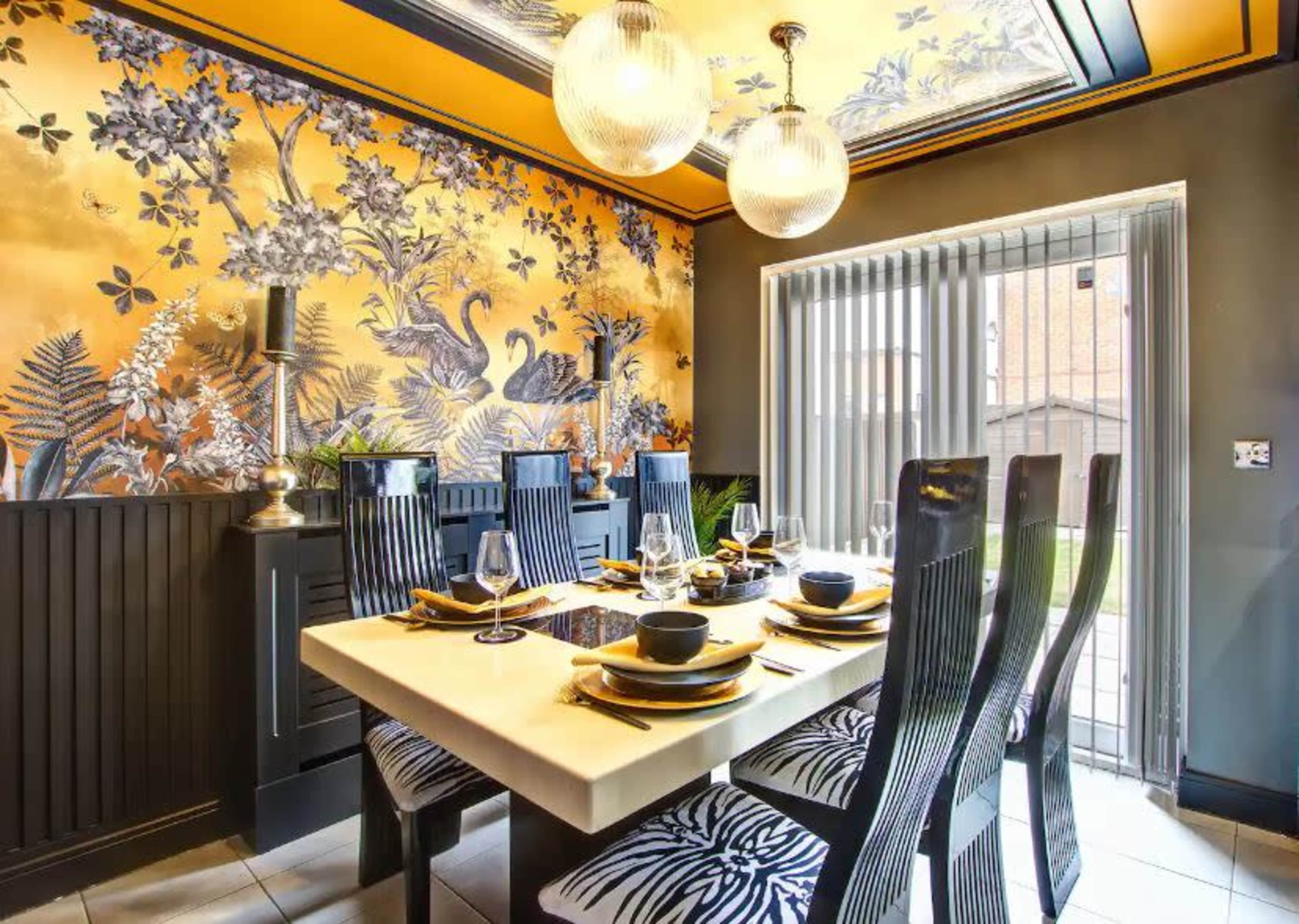 The dining room features a large white table set with black dishes and surrounded by chairs with zebra-patterned upholstery, against a backdrop of a gold and floral wallpaper.