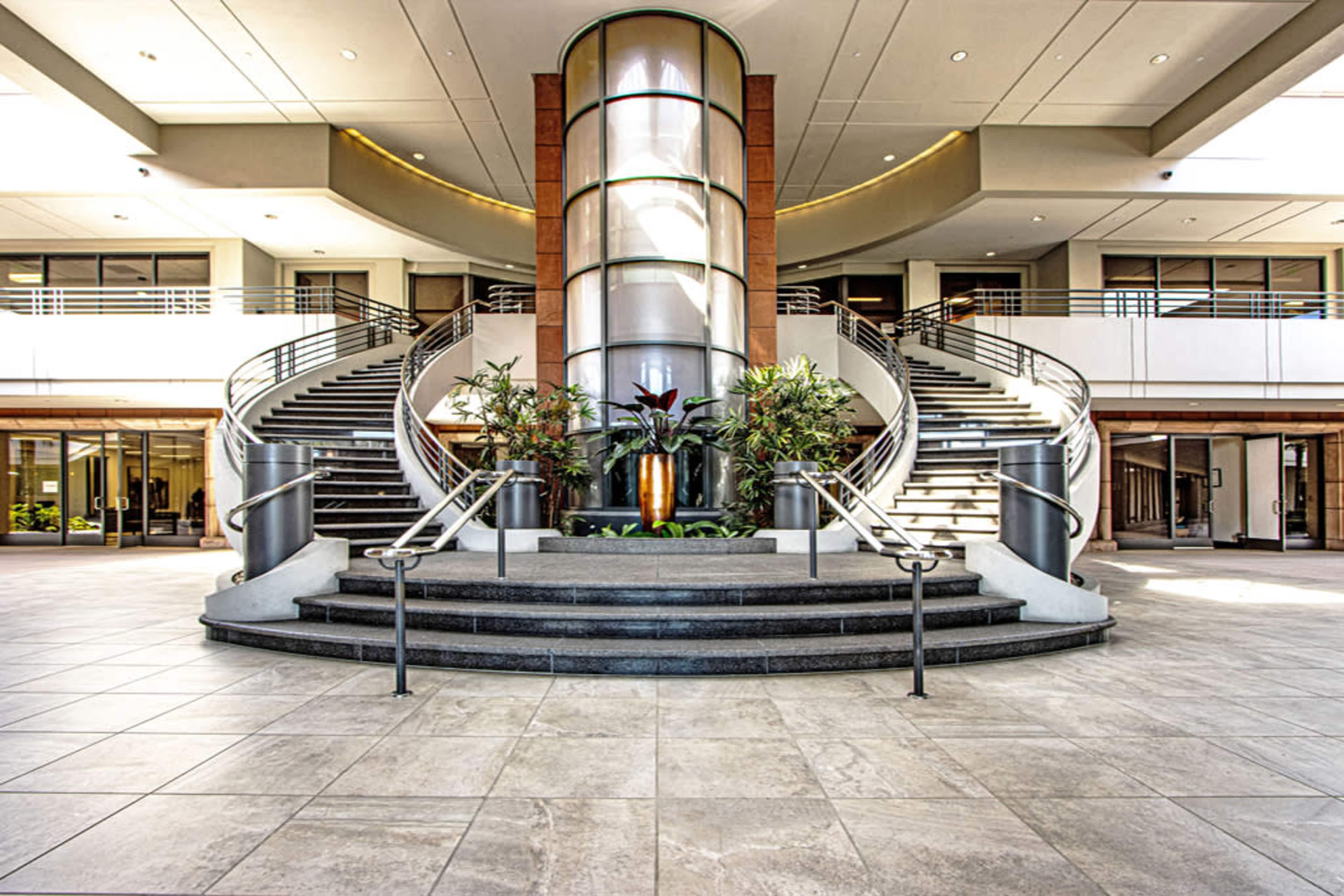 The image shows a spacious lobby featuring a circular staircase with surrounding greenery and an atrium-like structure.