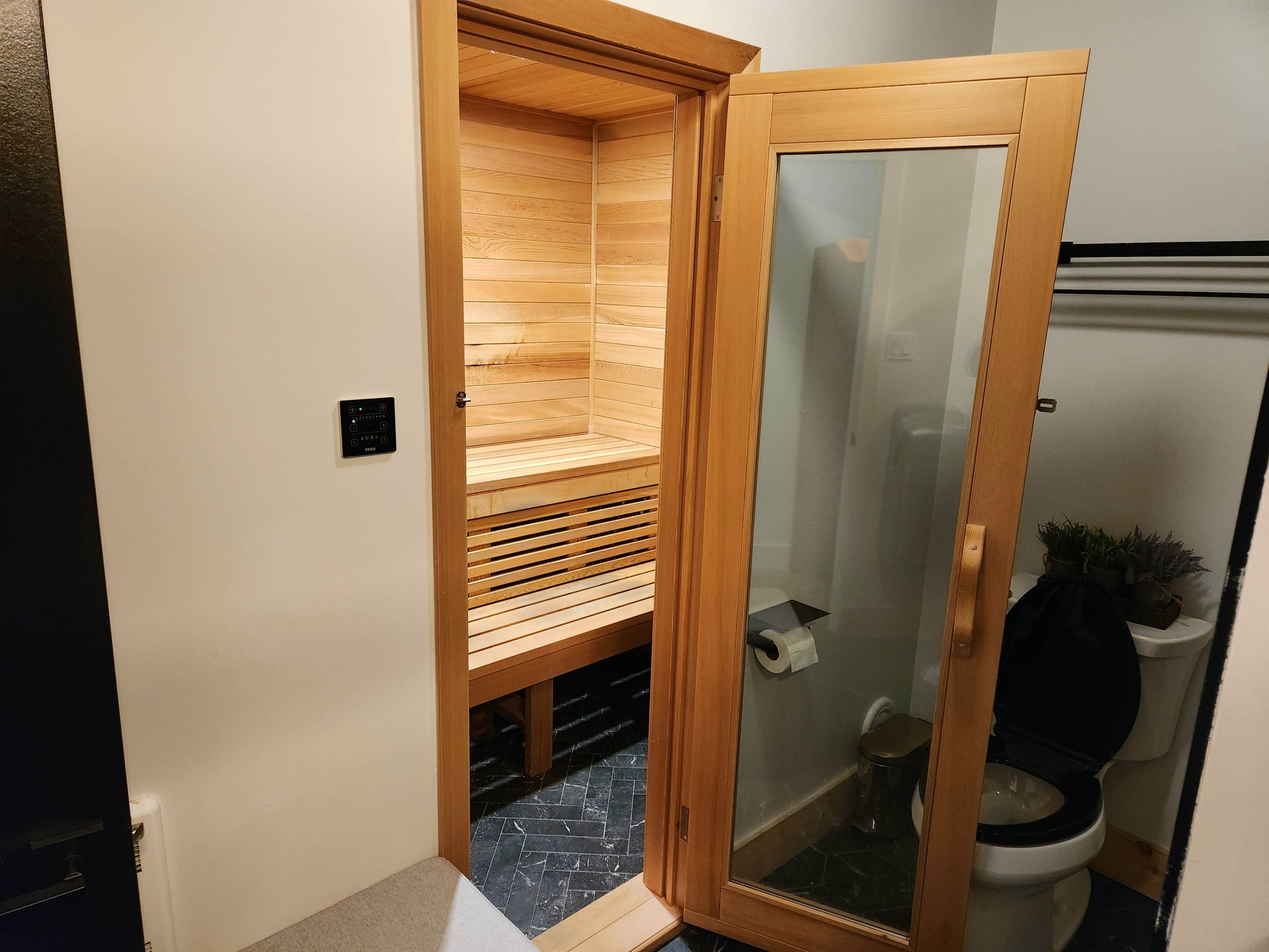 A wooden sauna is visible through an open door in a bathroom, adjacent to a toilet and a clear glass shower door.