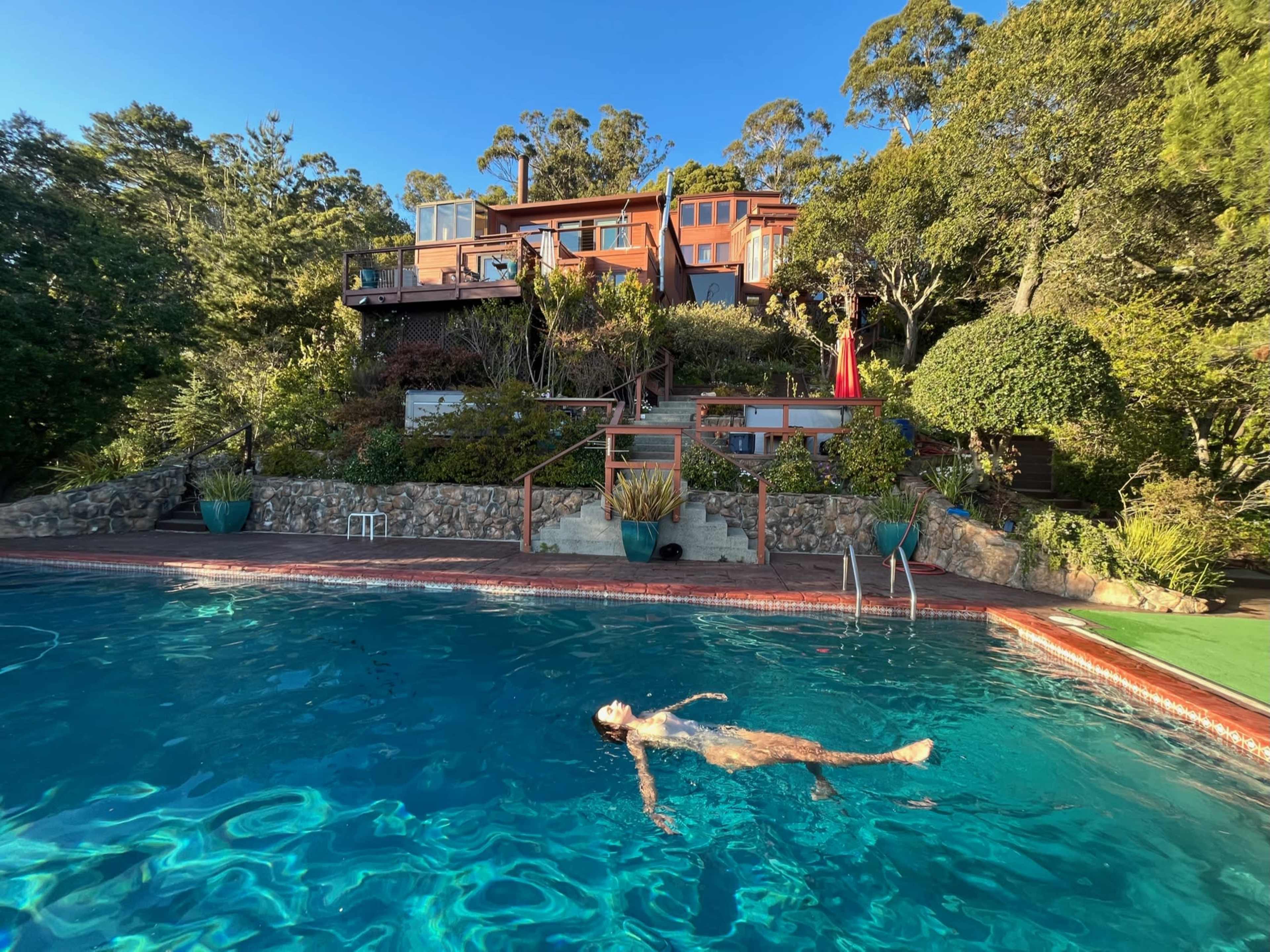 A person floats in a swimming pool in front of a multi-level house surrounded by trees and greenery.