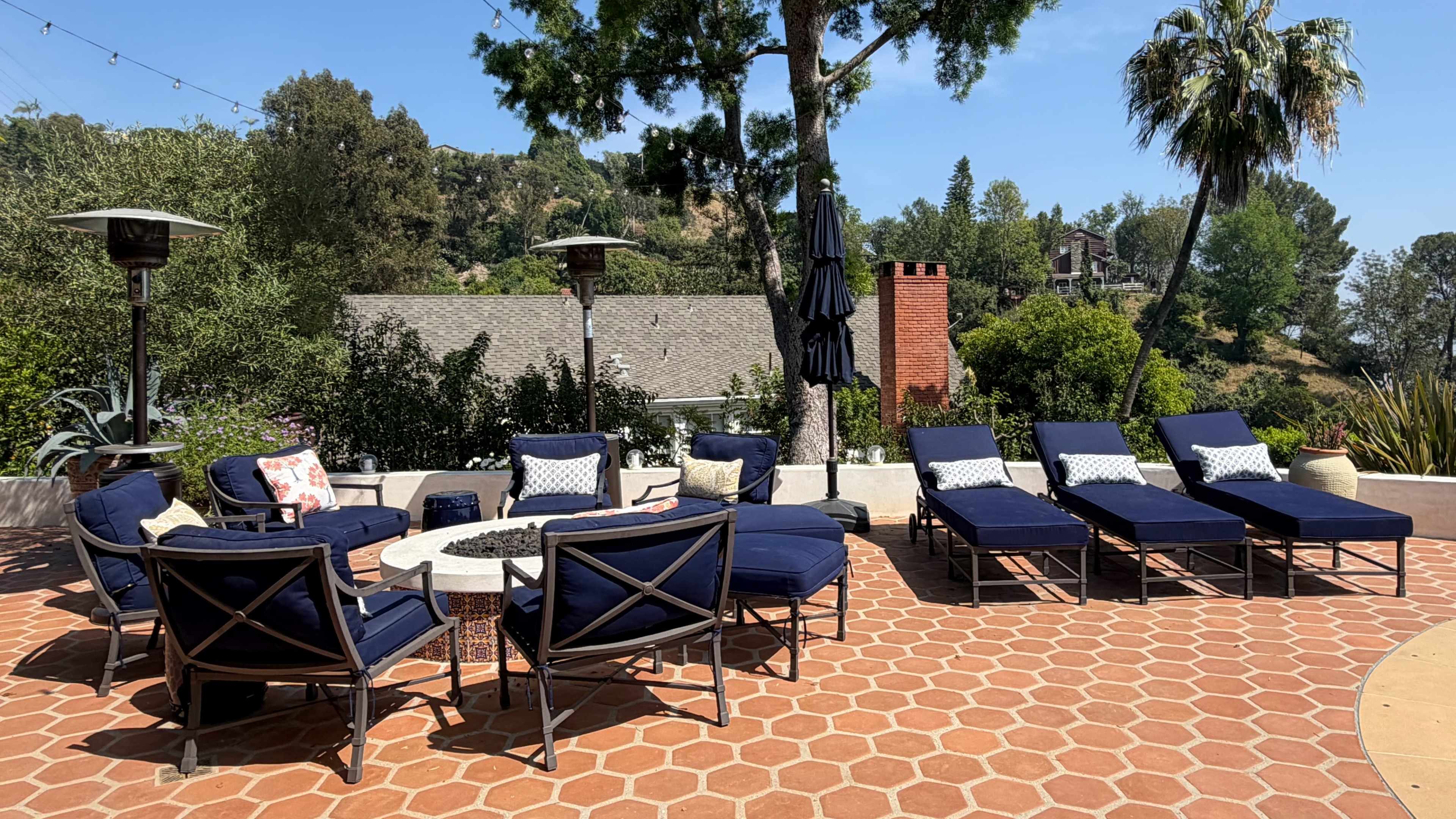 The image shows a patio area with several navy blue lounge chairs arranged around a circular fire pit, surrounded by greenery and a few trees.
