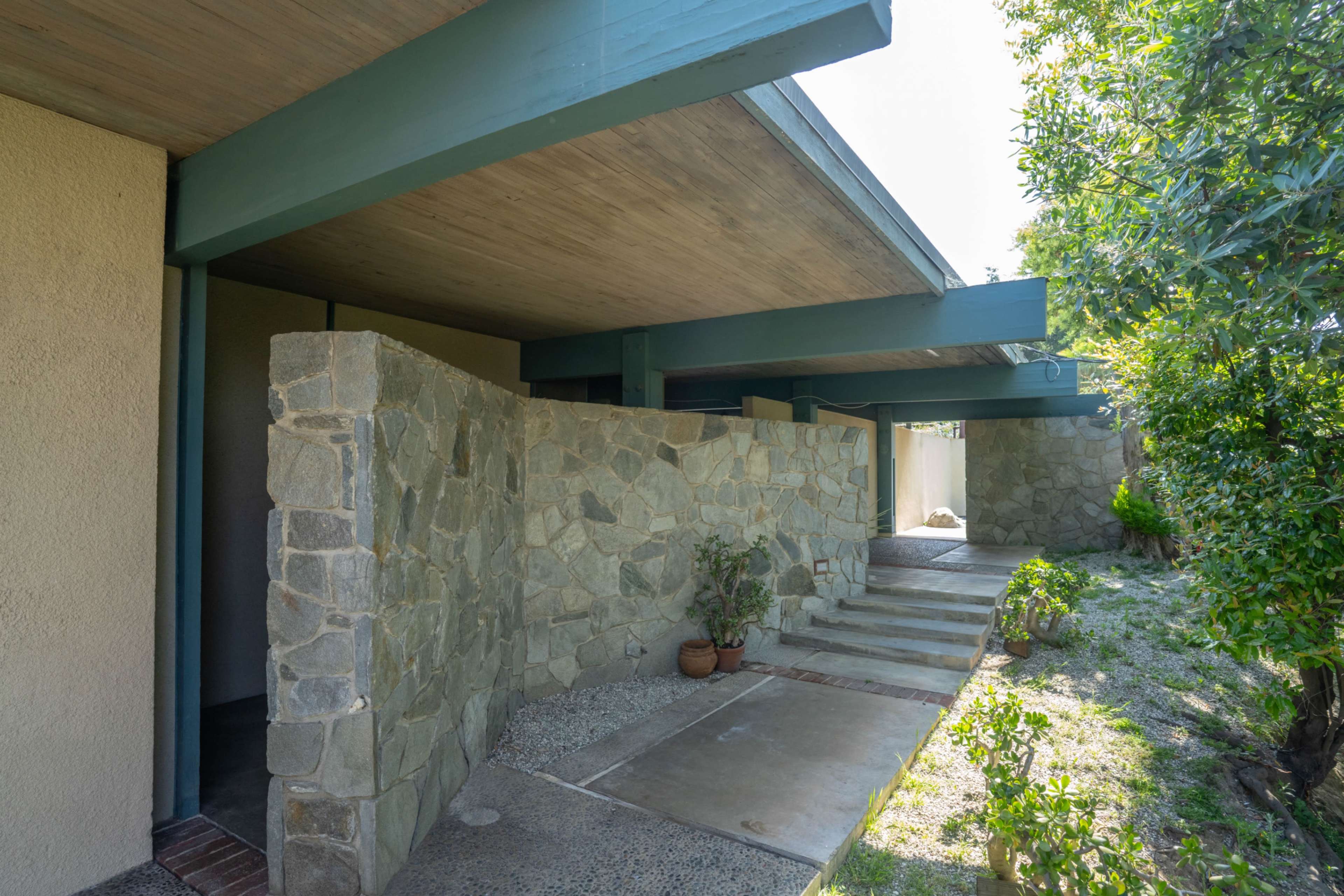 The image shows a modern architectural structure with a stone wall, concrete pathway, and greenery surrounding the entrance.