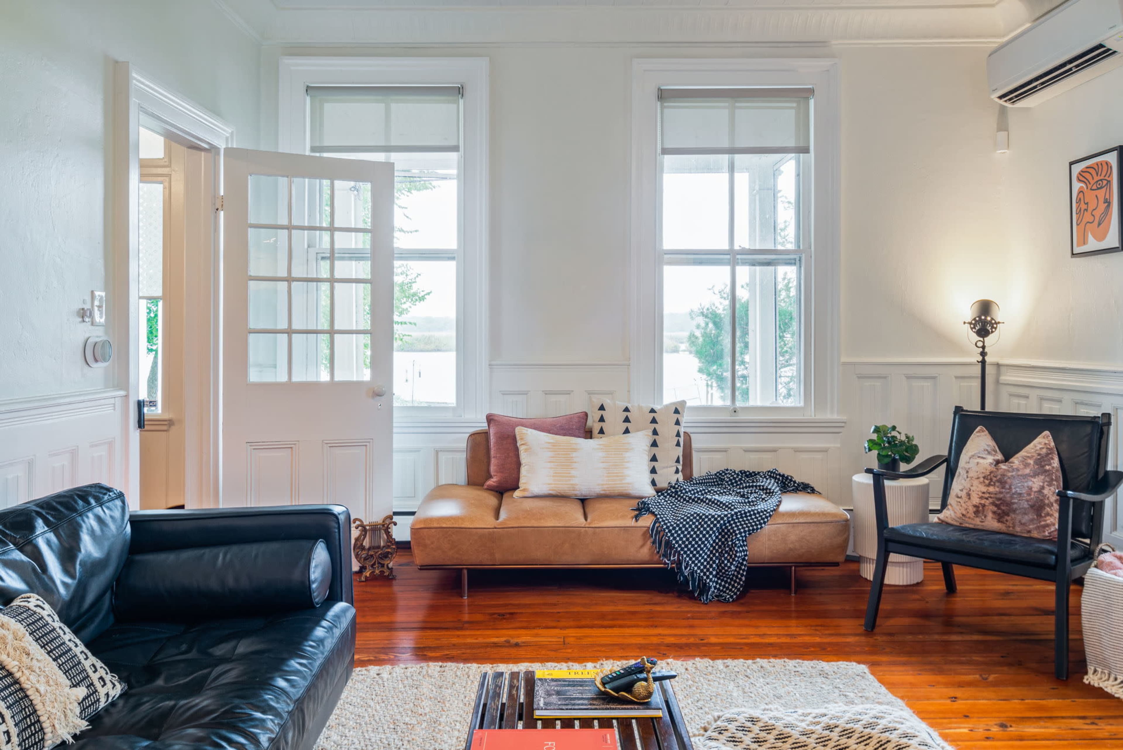 The image shows a living room with hardwood flooring, featuring a black leather couch, a tan sofa with decorative cushions, and large windows allowing natural light to enter.