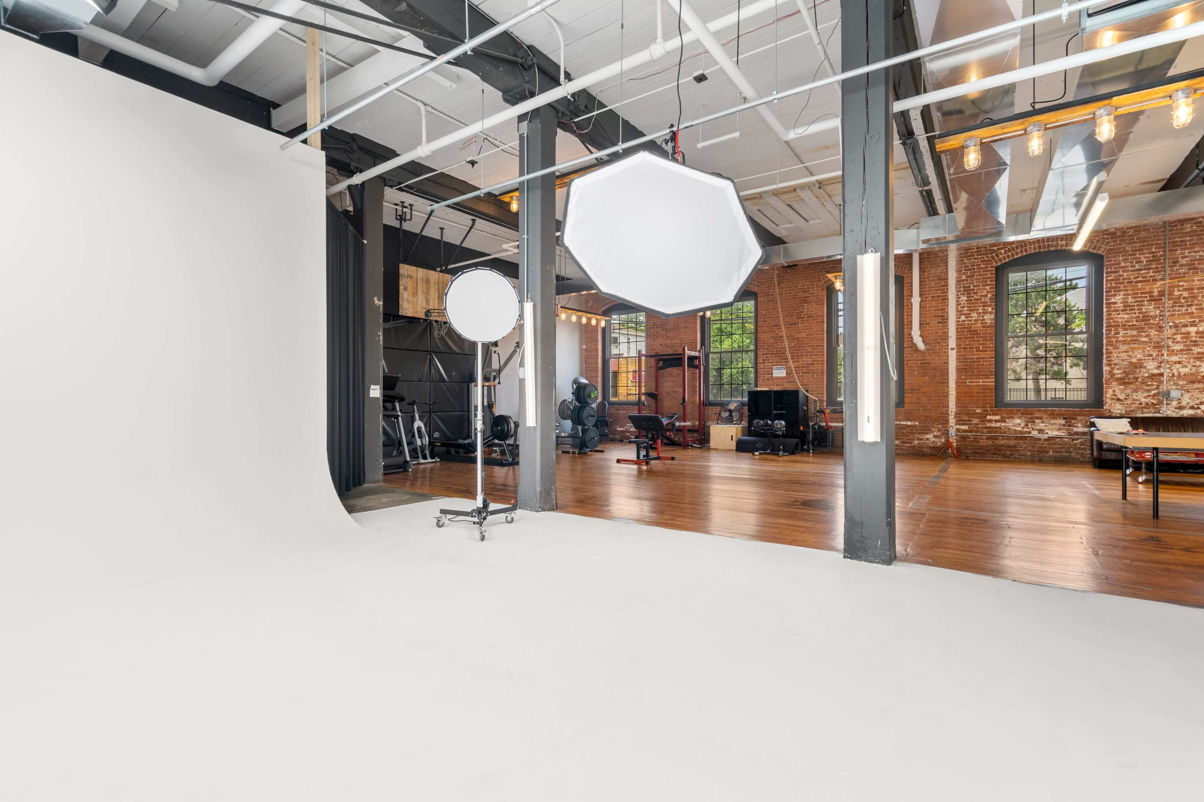 A spacious photography studio with a white backdrop, a large light modifier, and exposed brick walls, showcasing wooden flooring and various equipment in the background.