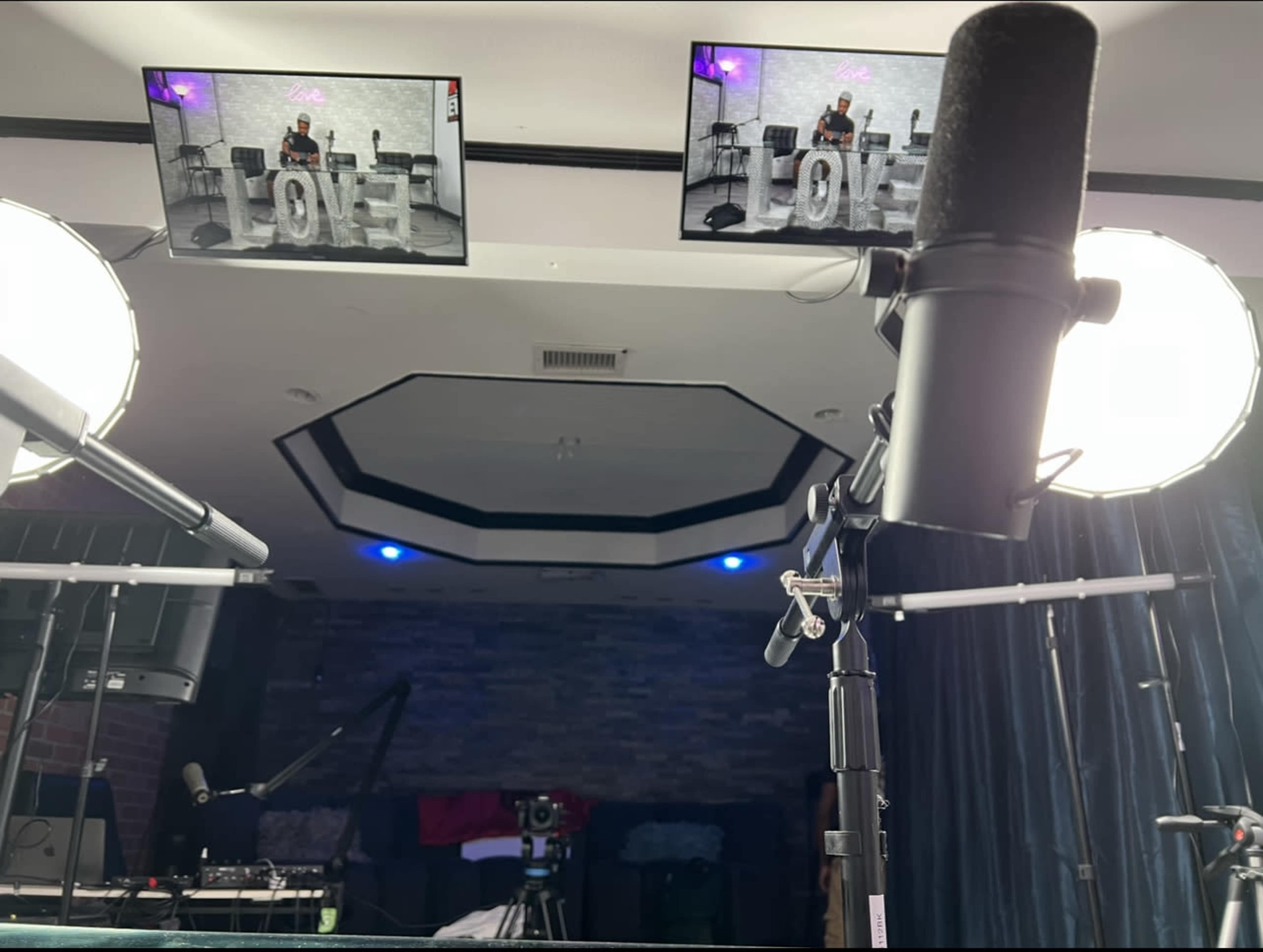 The image shows a recording setup with two overhead screens displaying the word "LOVE," multiple microphones, and softbox lights in a room with a dark backdrop.