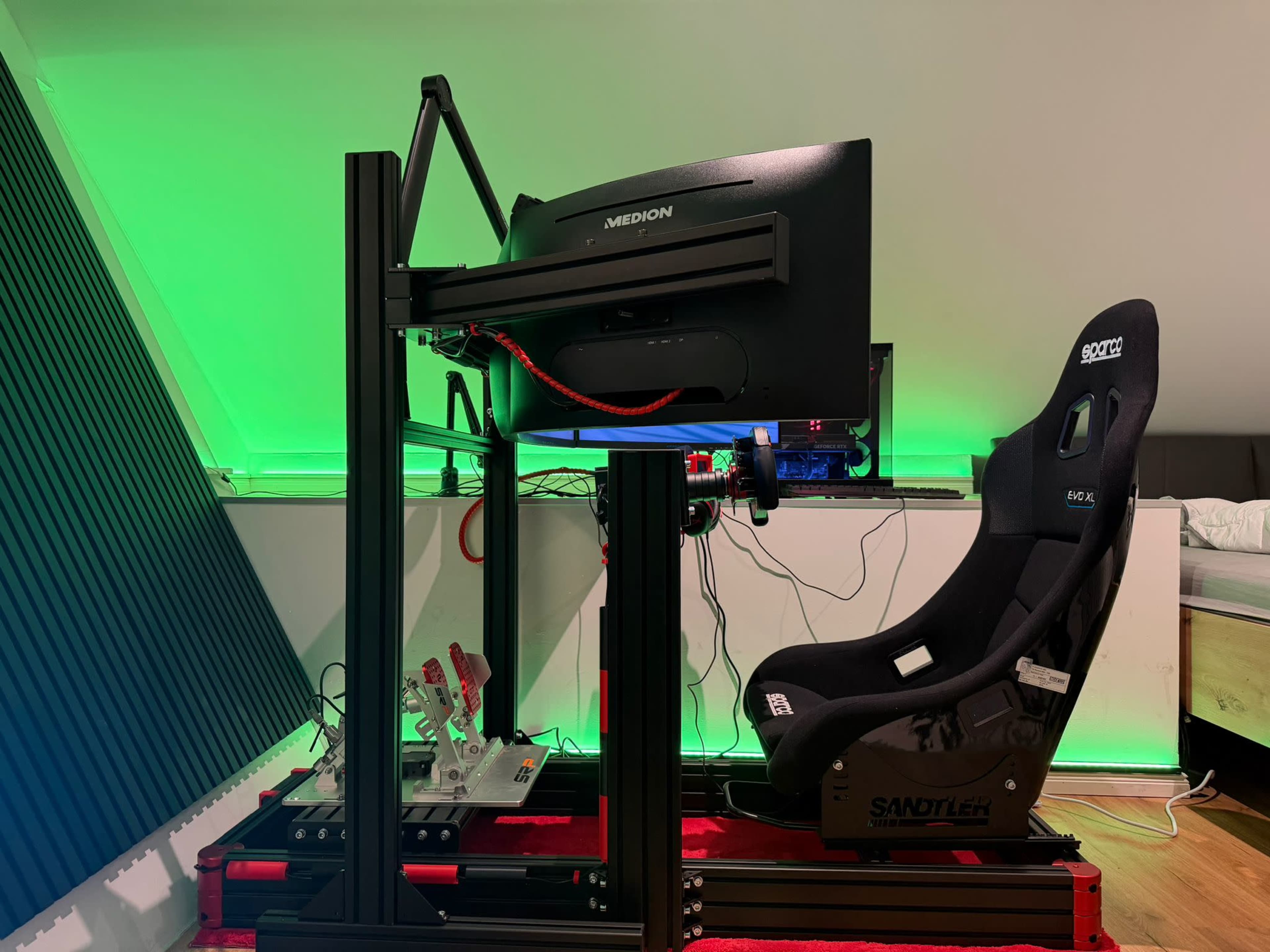 The image shows a racing simulator setup with a black gaming chair, a monitor mounted on a stand, and pedals arranged under the chair, all positioned in a room with green ambient lighting.