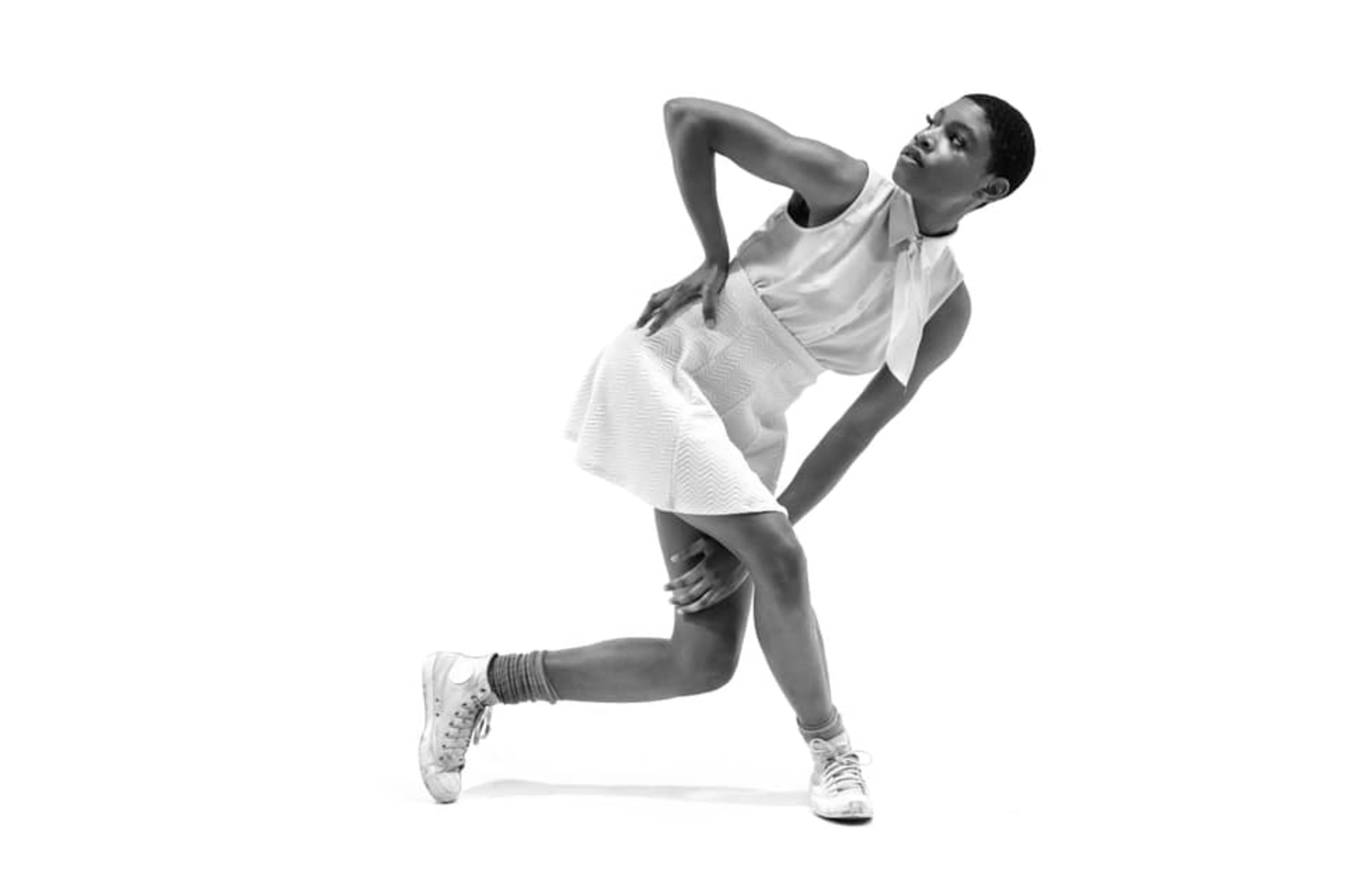 A person in a white dress and sneakers poses with one hand on their hip and the other on their knee against a plain white background.