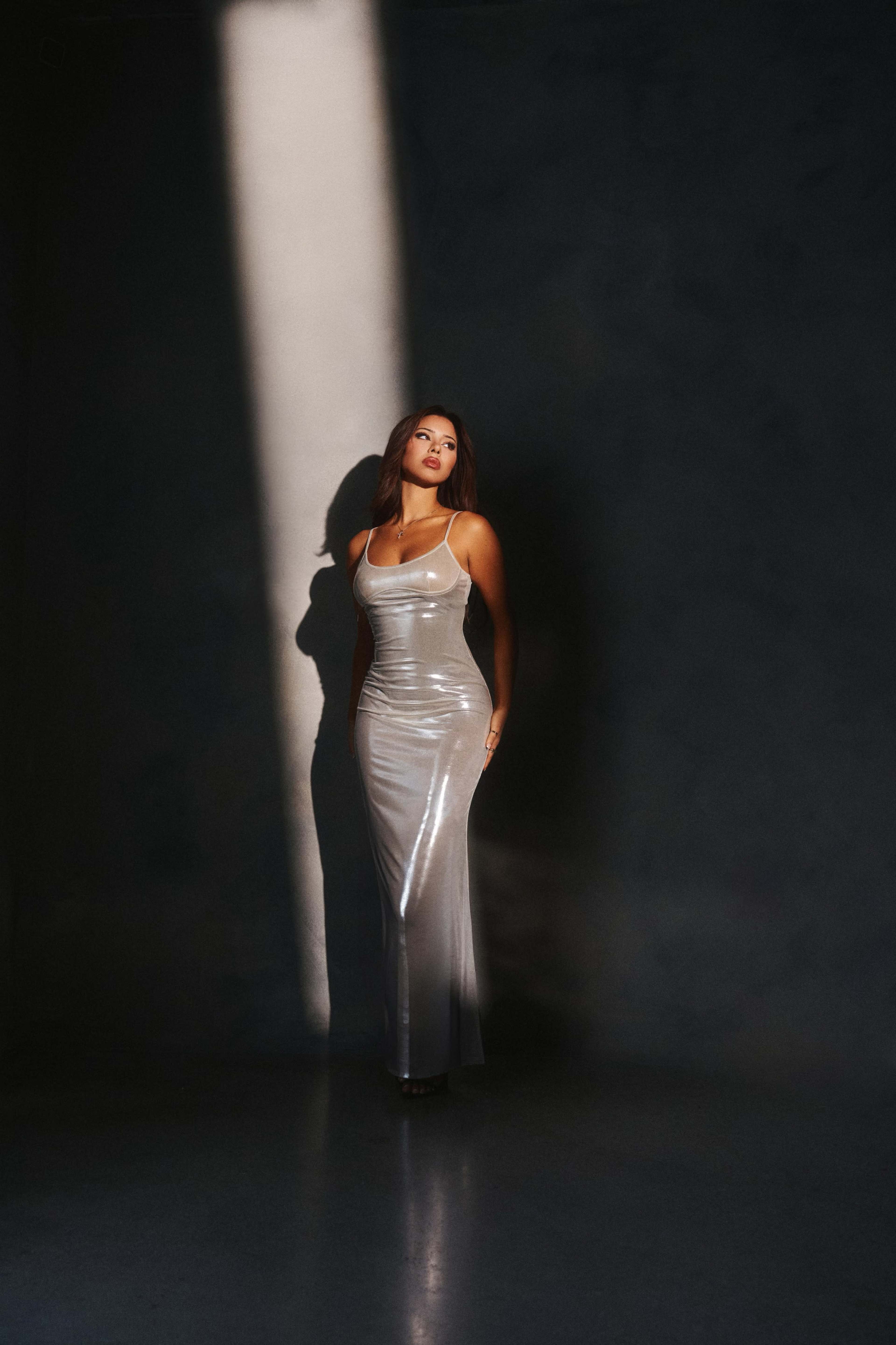 A woman in a shiny silver gown stands in a dark room, illuminated by a beam of light.