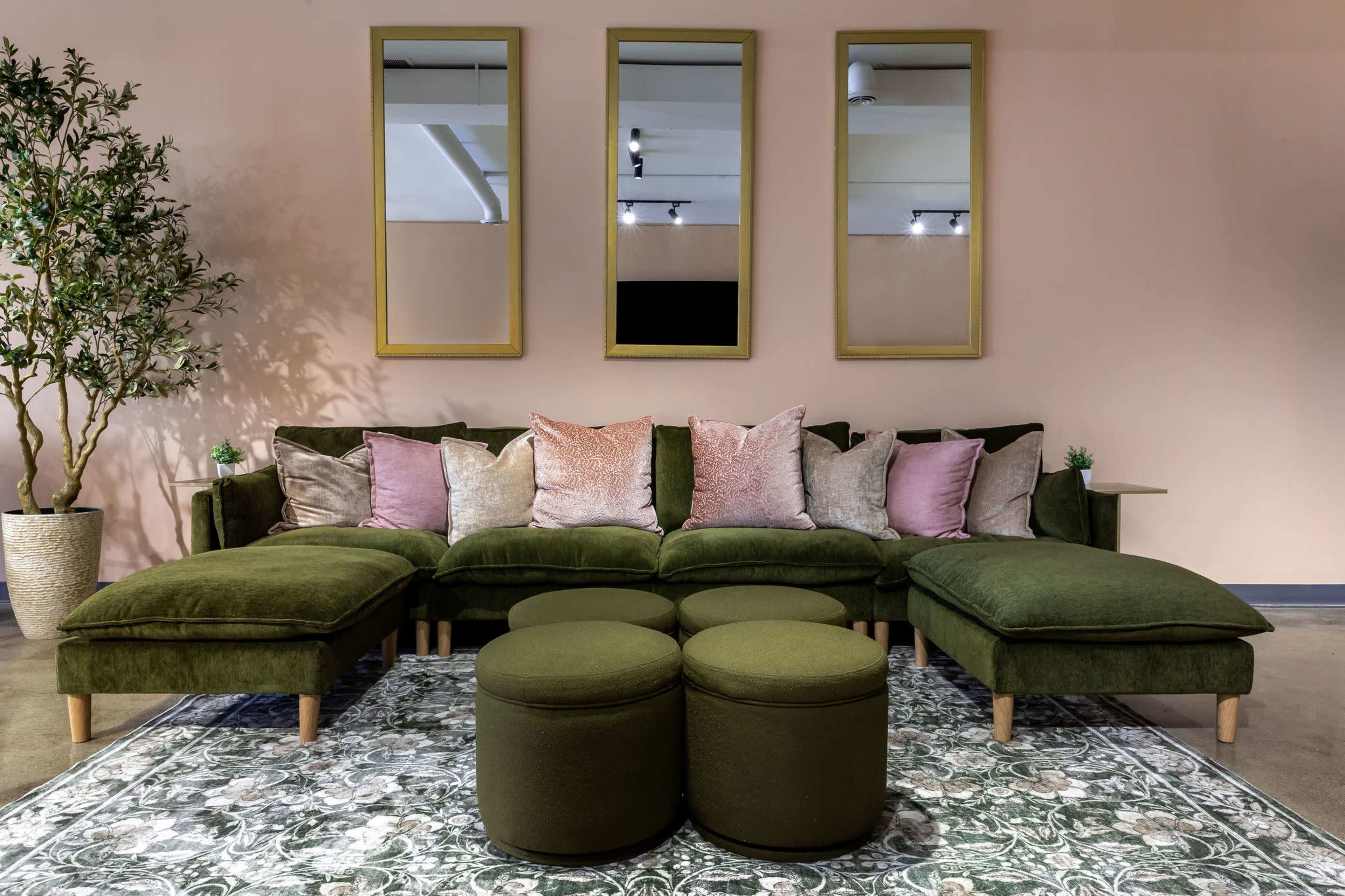 A sectional sofa with various throw pillows is positioned in front of three mirrored frames against a pastel wall, accompanied by a small potted plant and round ottomans on a patterned rug.
