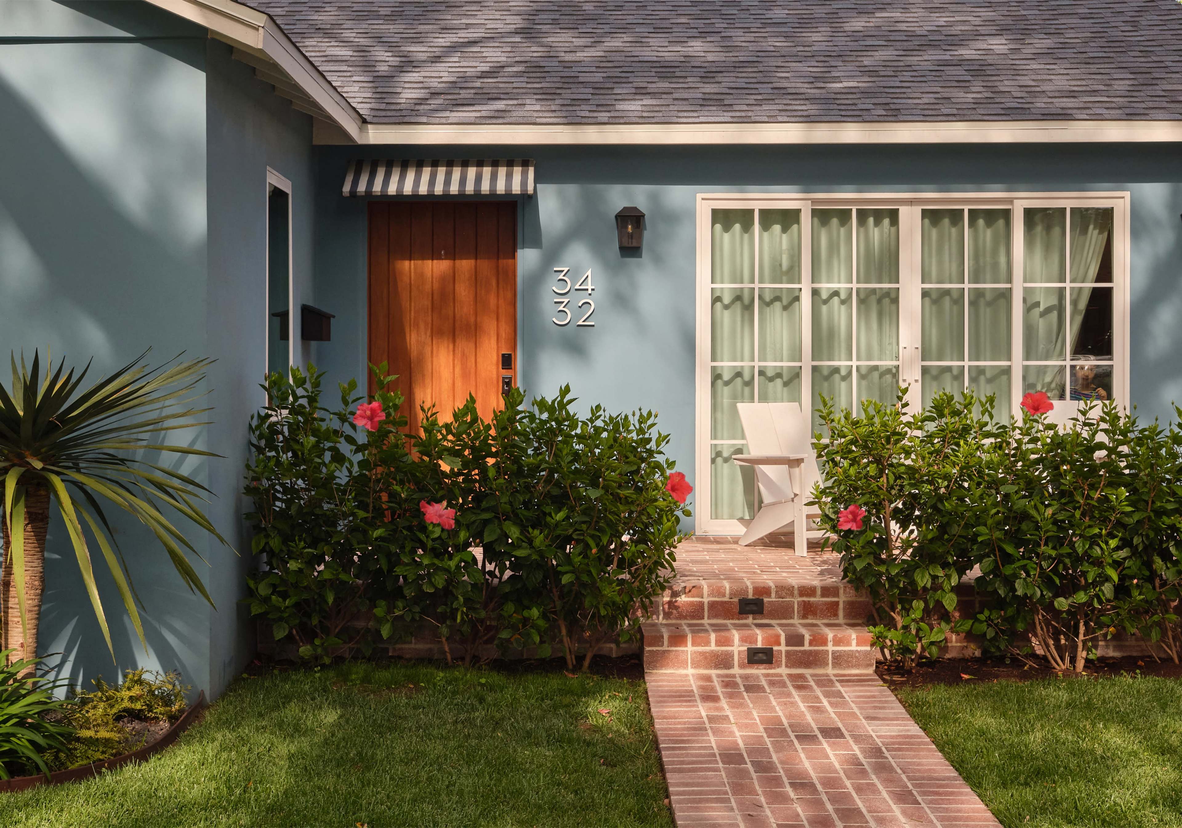 The image shows a well-maintained home facade with a blue exterior, a brick pathway, and green shrubbery, featuring a wooden door and a large window with sheer curtains.
