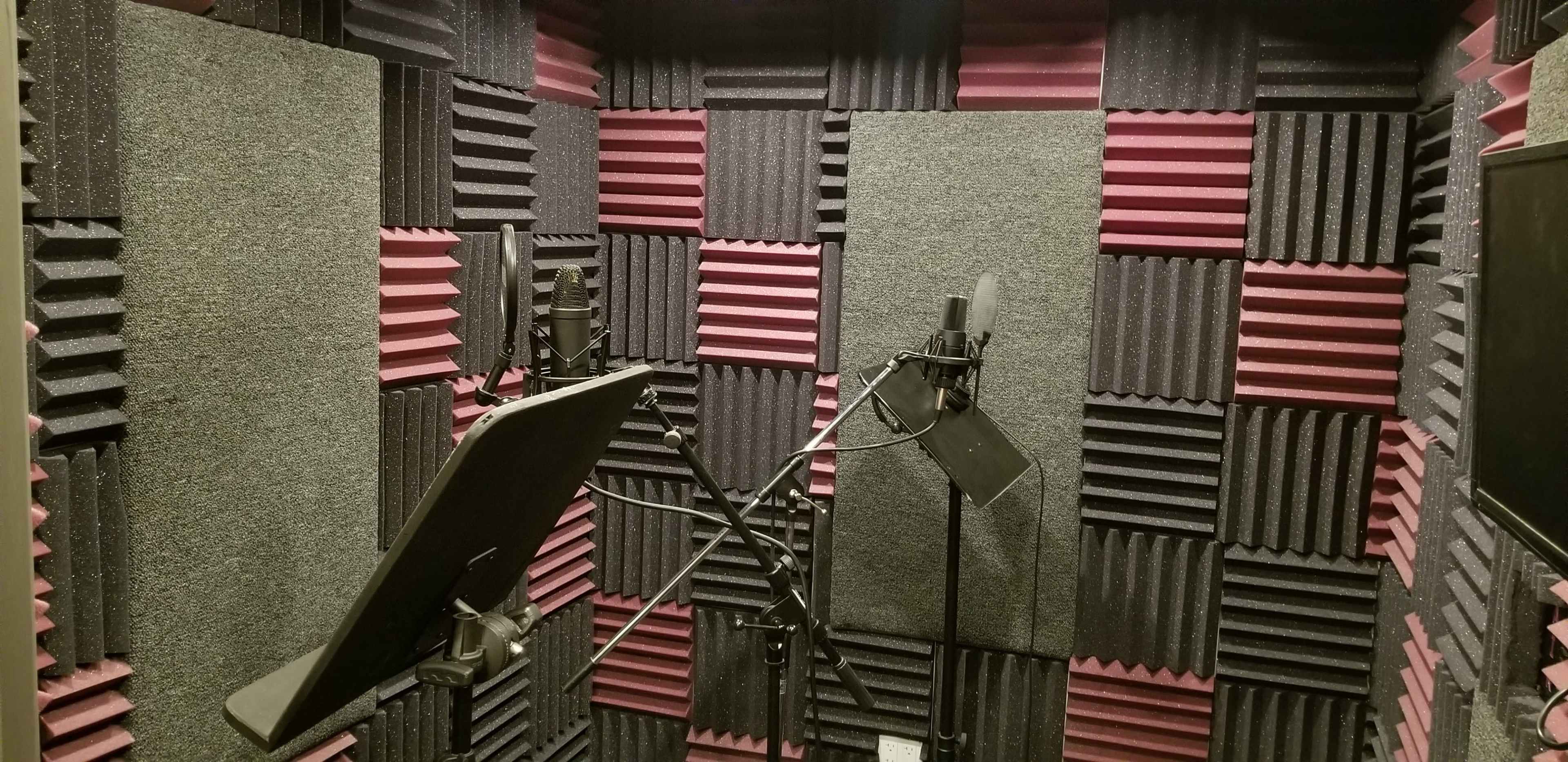 A soundproof recording booth with acoustic foam panels in black and pink, equipped with two microphones on stands and a music stand.