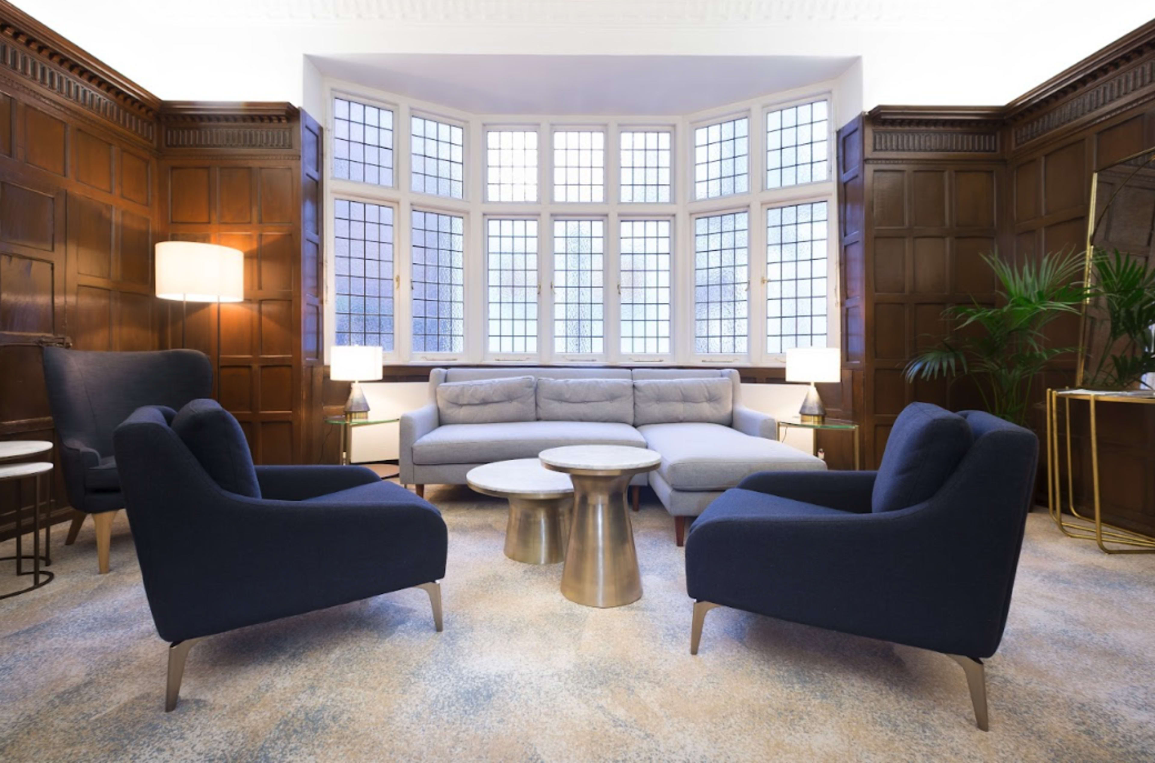 A modern sitting area features a gray sofa flanked by two dark blue chairs, with a round table in the center and large windows illuminating the space.