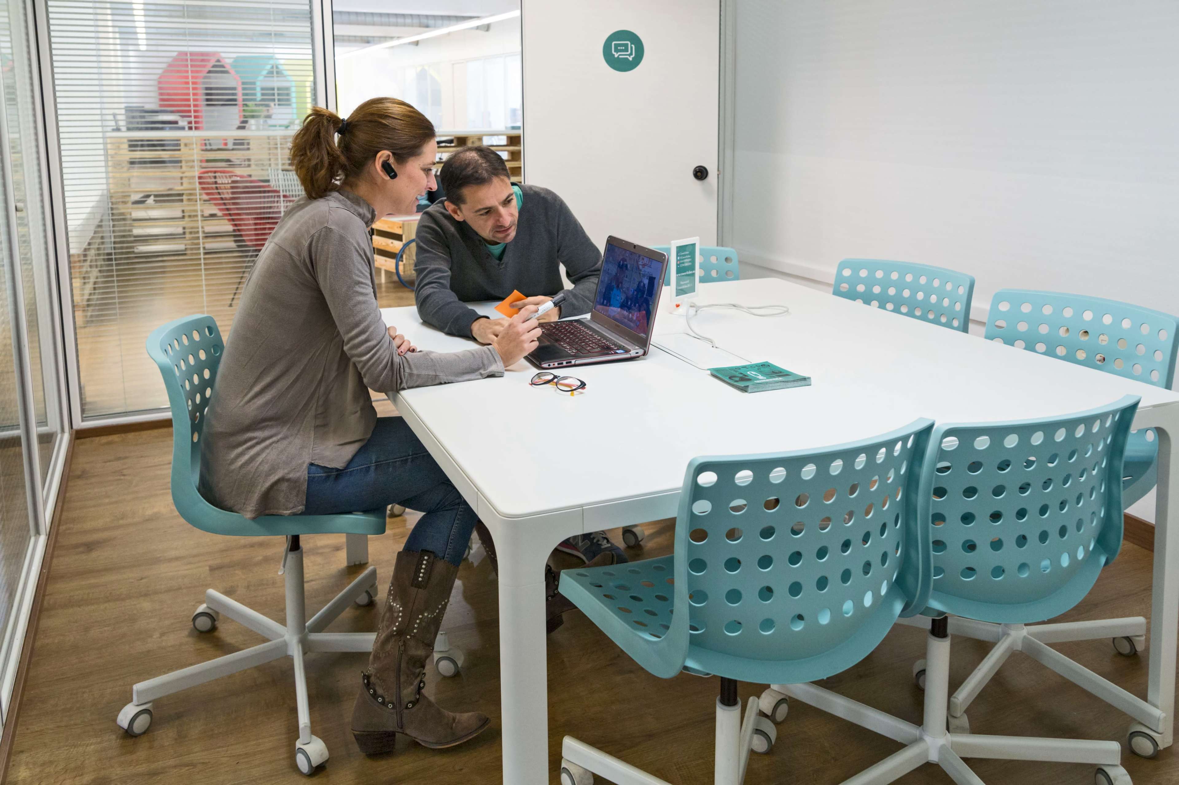 A woman and a man review a laptop together at a white table in a modern meeting room.