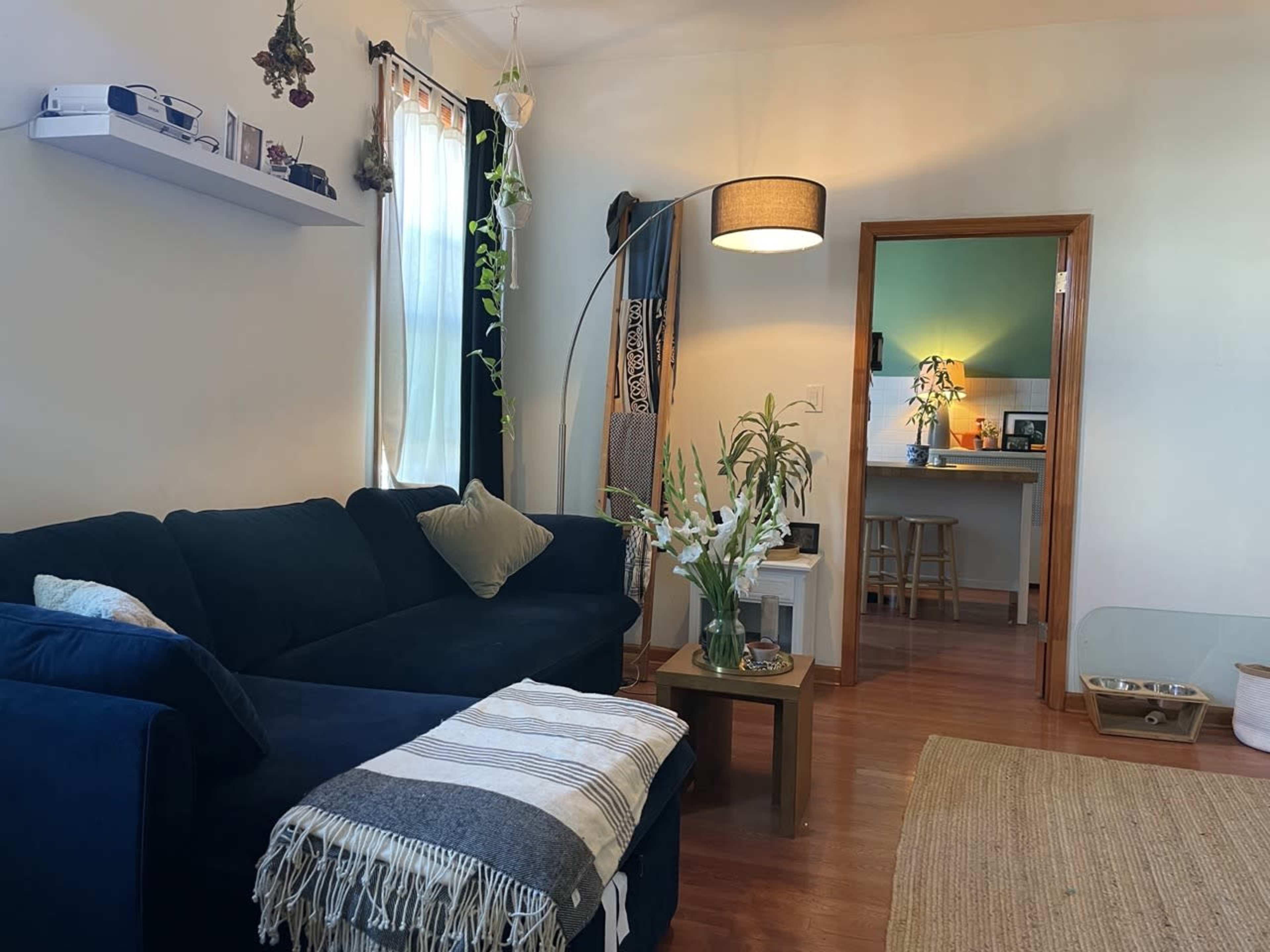 A cozy living room with a dark blue sofa, a small coffee table, a lamp, and a view of a kitchen area through an open door.