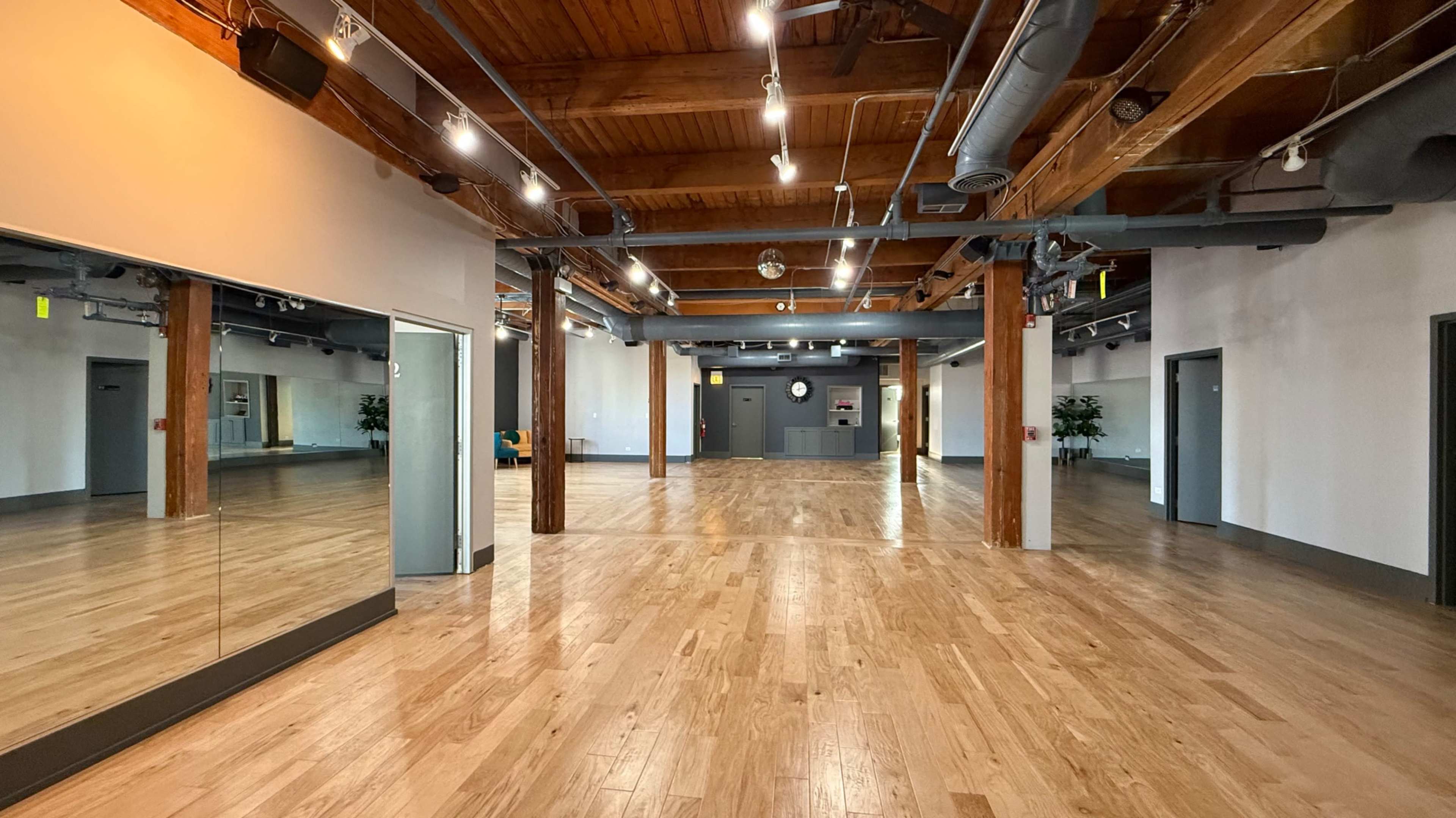 The image shows a spacious, empty room with wooden floors, exposed beams, and large mirrors on one wall.