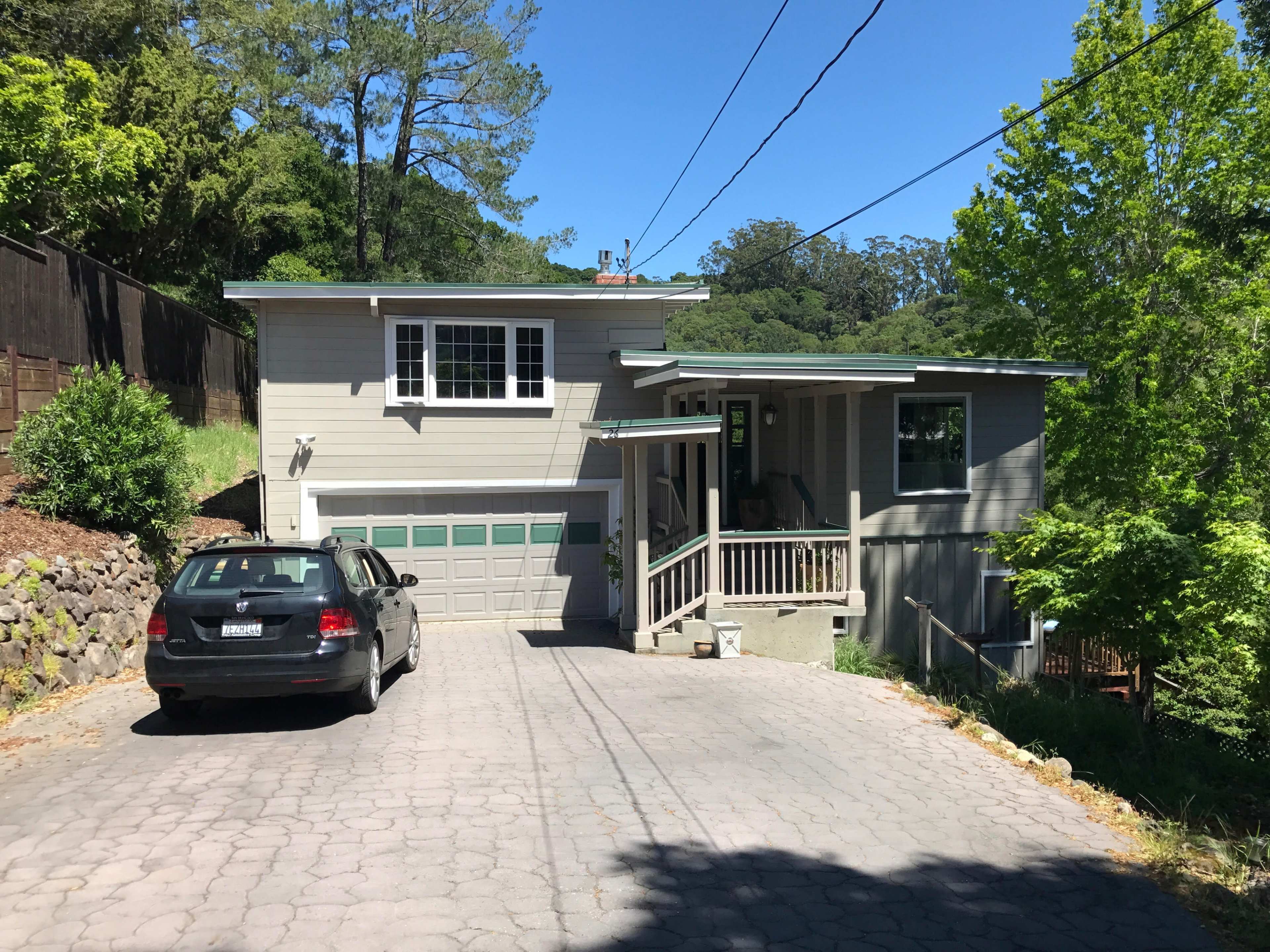 A two-story gray house with a garage is situated on a sloped driveway surrounded by trees.