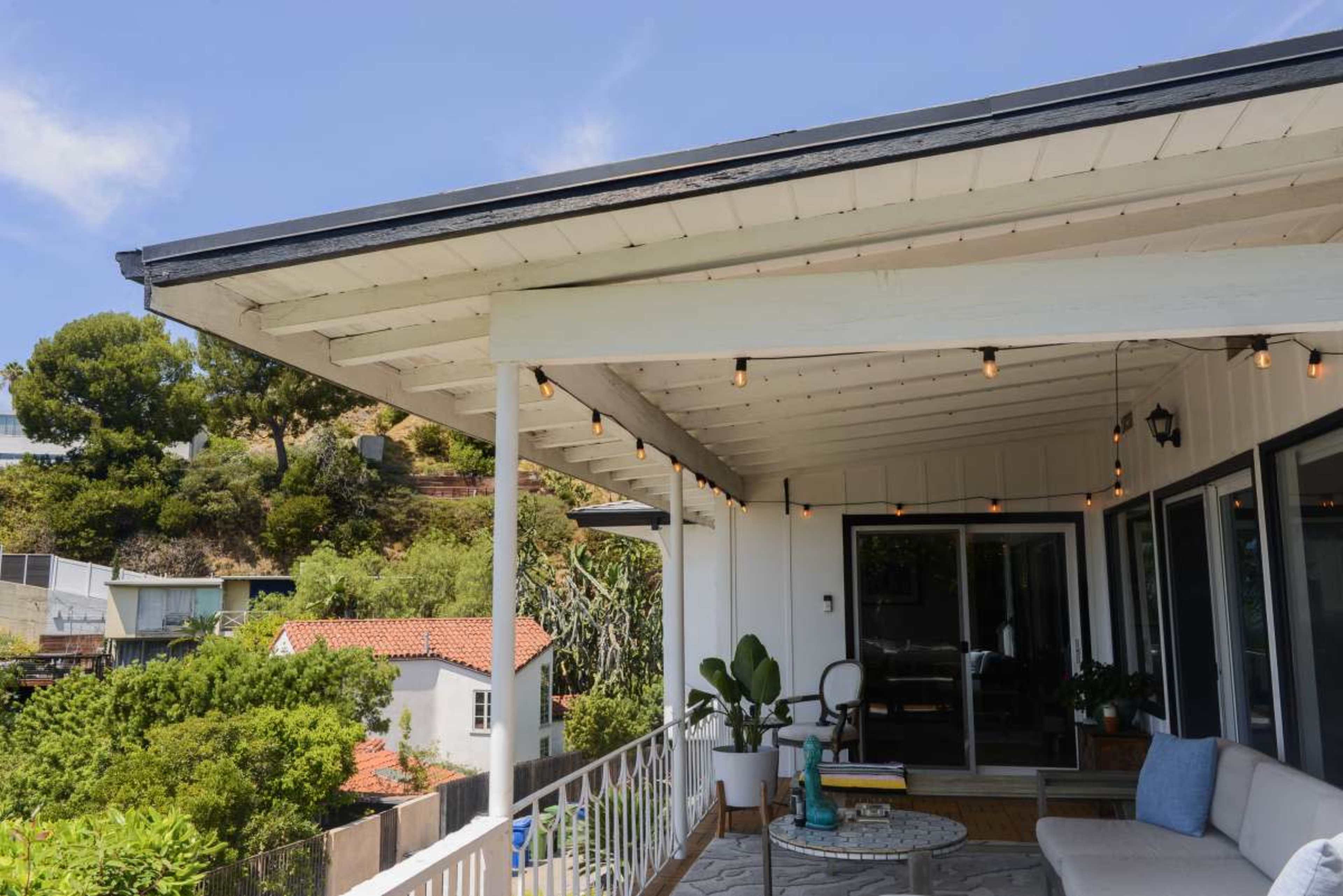 The image shows a modern outdoor patio area of a house with a white overhang and string lights, overlooking greenery and neighboring homes.