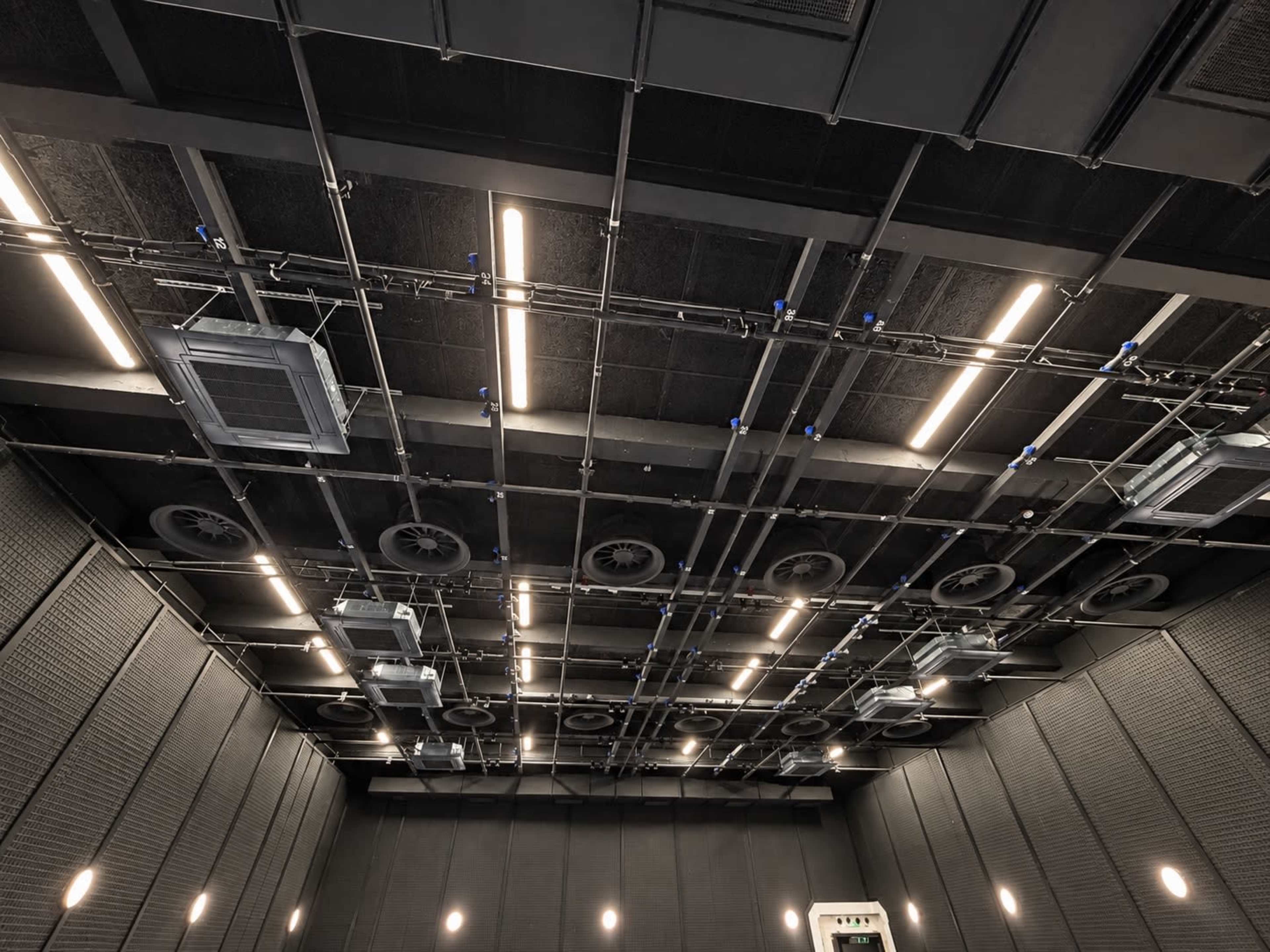 The image shows a large, industrial ceiling with exposed ventilation systems, lights, and structural supports in a dark room.