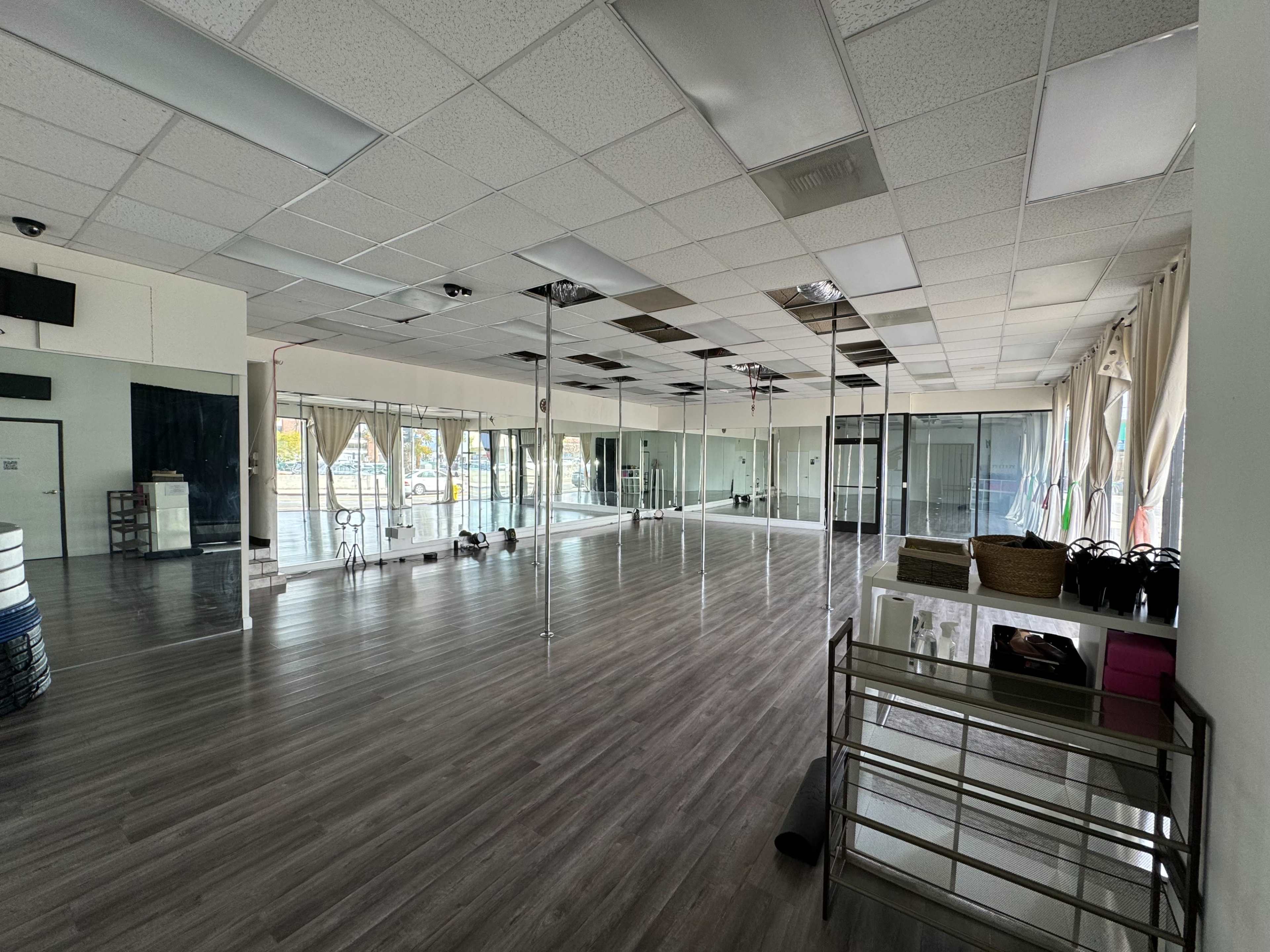 An empty dance studio with mirrors lining one wall, wooden flooring, and various exercise equipment in the corner.