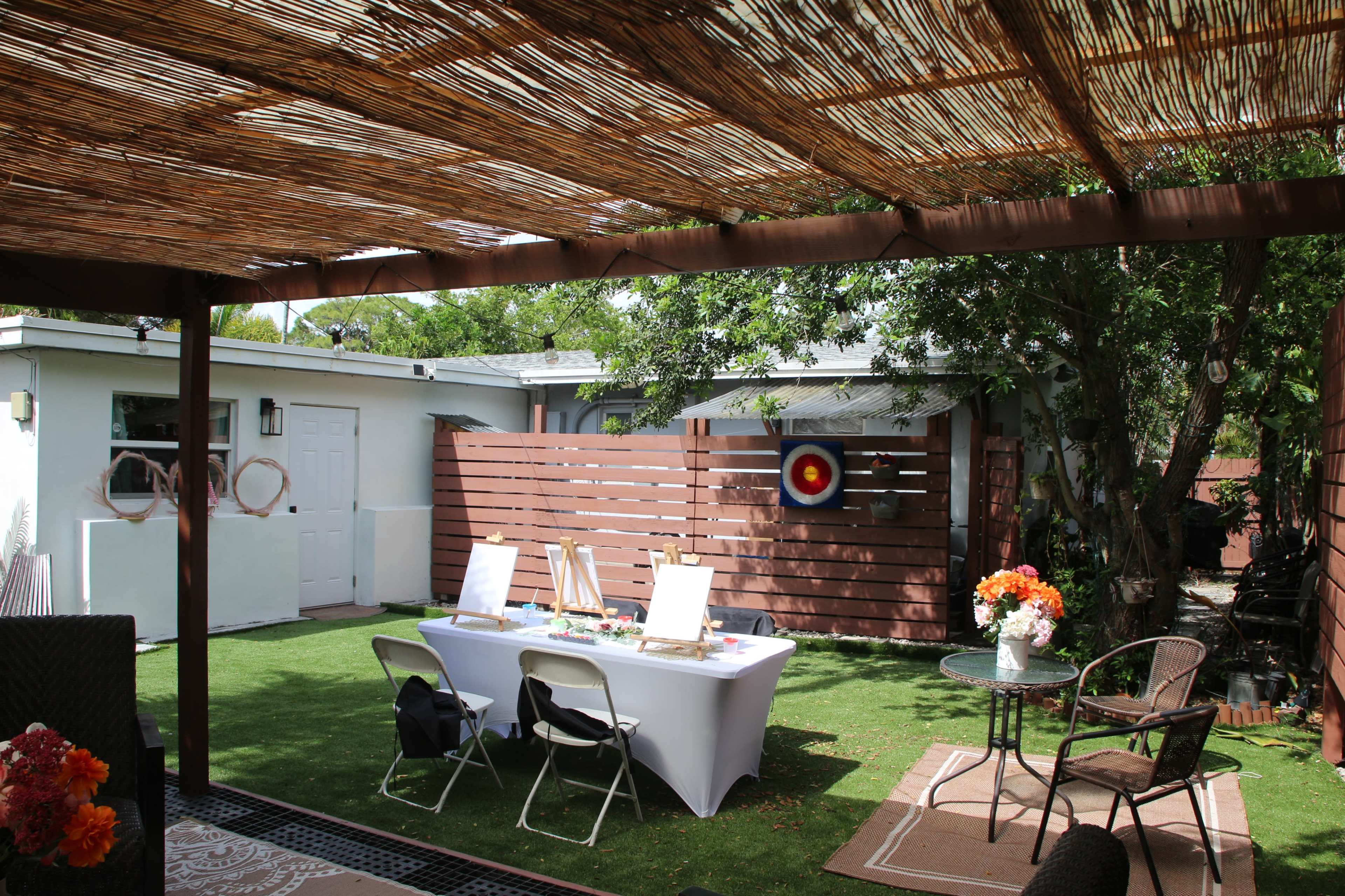 A patio featuring a table set with easels and art supplies, surrounded by greenery and a wall with a target painting.