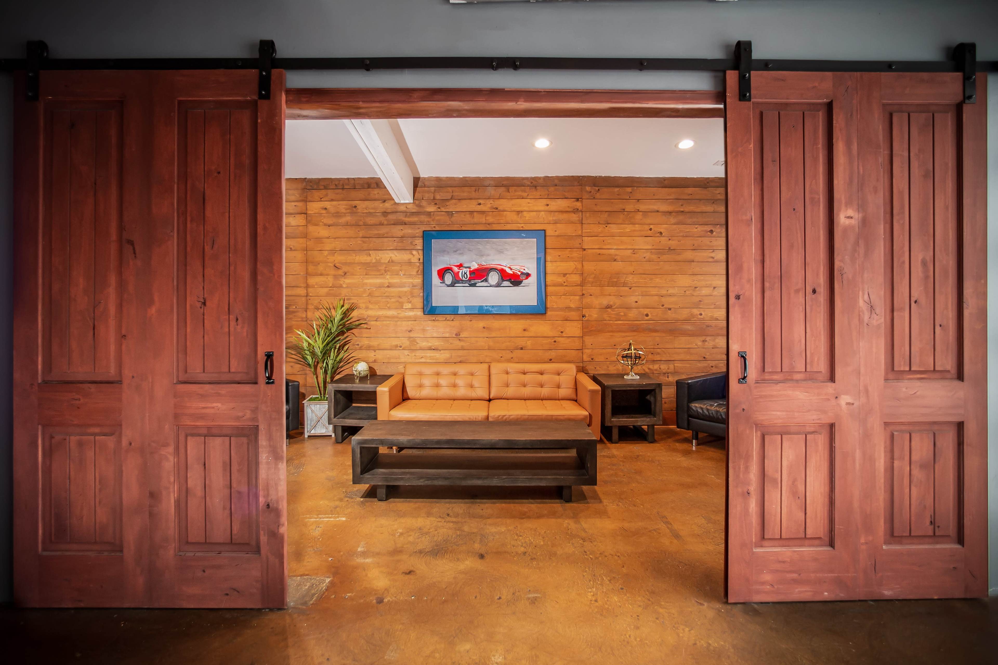 The image shows a room with wooden sliding doors revealing a seating area that features a tan sofa, a dark coffee table, and wooden walls adorned with a framed picture of a red sports car.