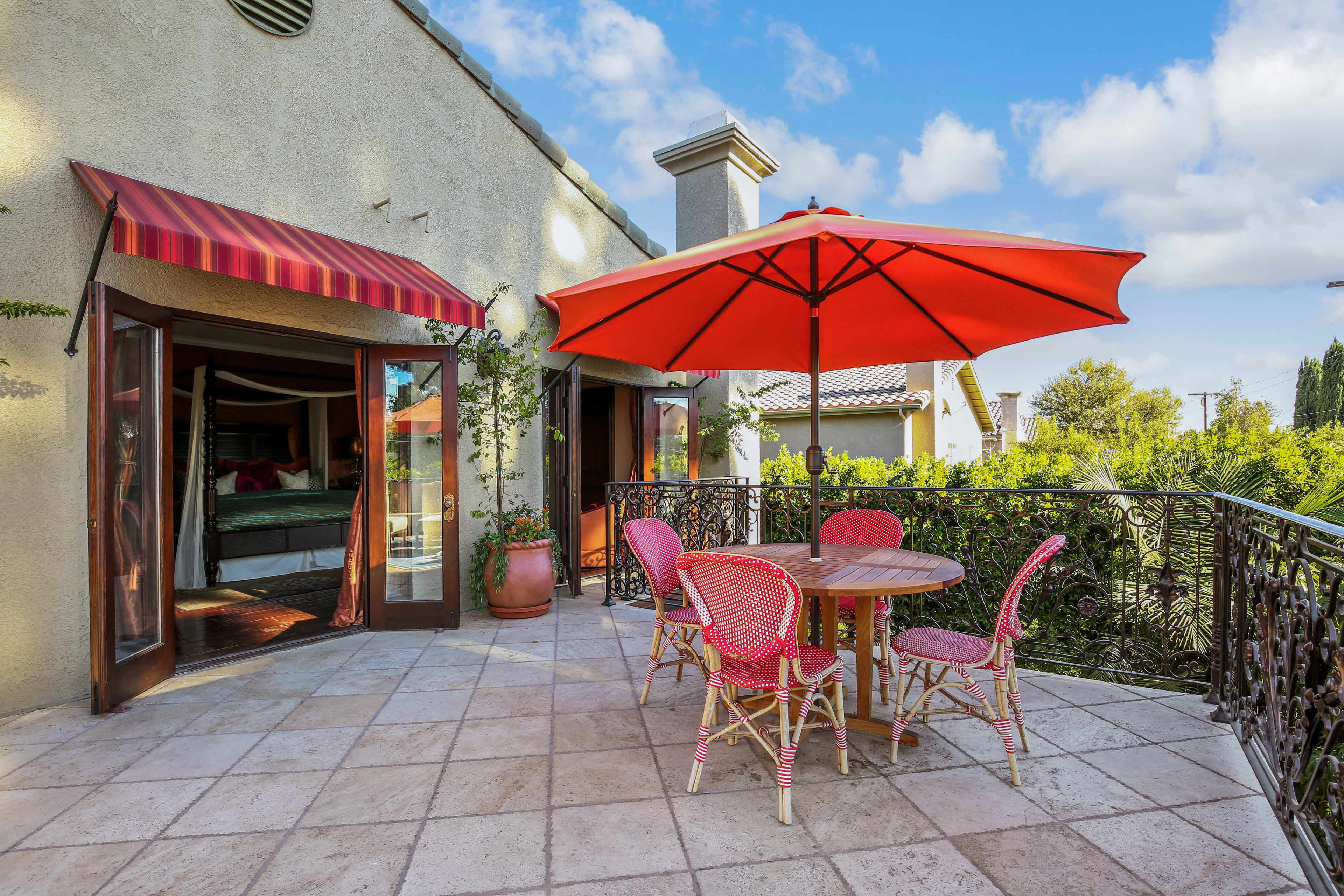 A patio features a round table surrounded by four pink chairs and a large red umbrella, set against a backdrop of greenery and a blue sky.