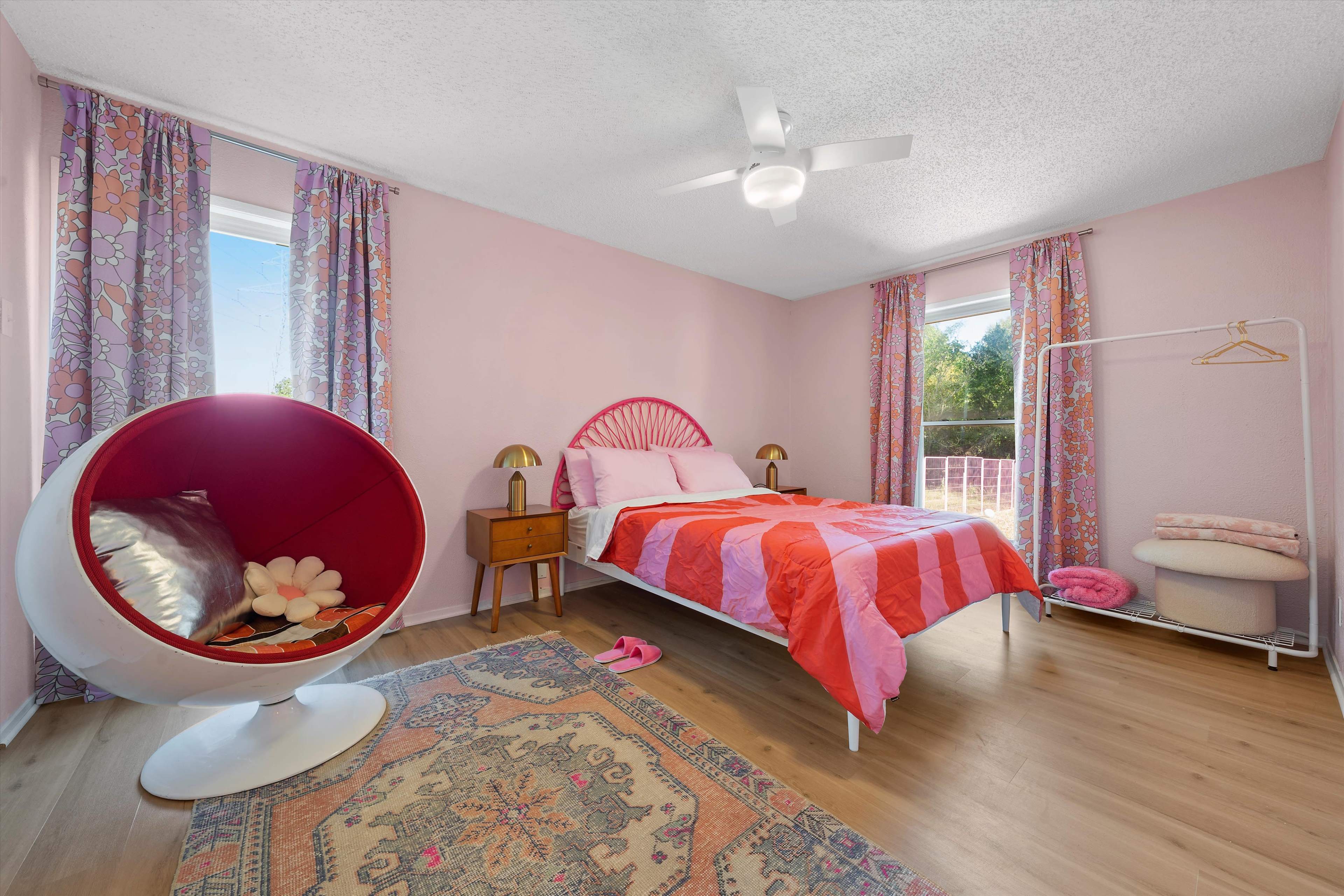 A bedroom featuring a pink color scheme, a round chair with red and white accents, a bed with a striped pink and red blanket, and large windows with floral curtains.
