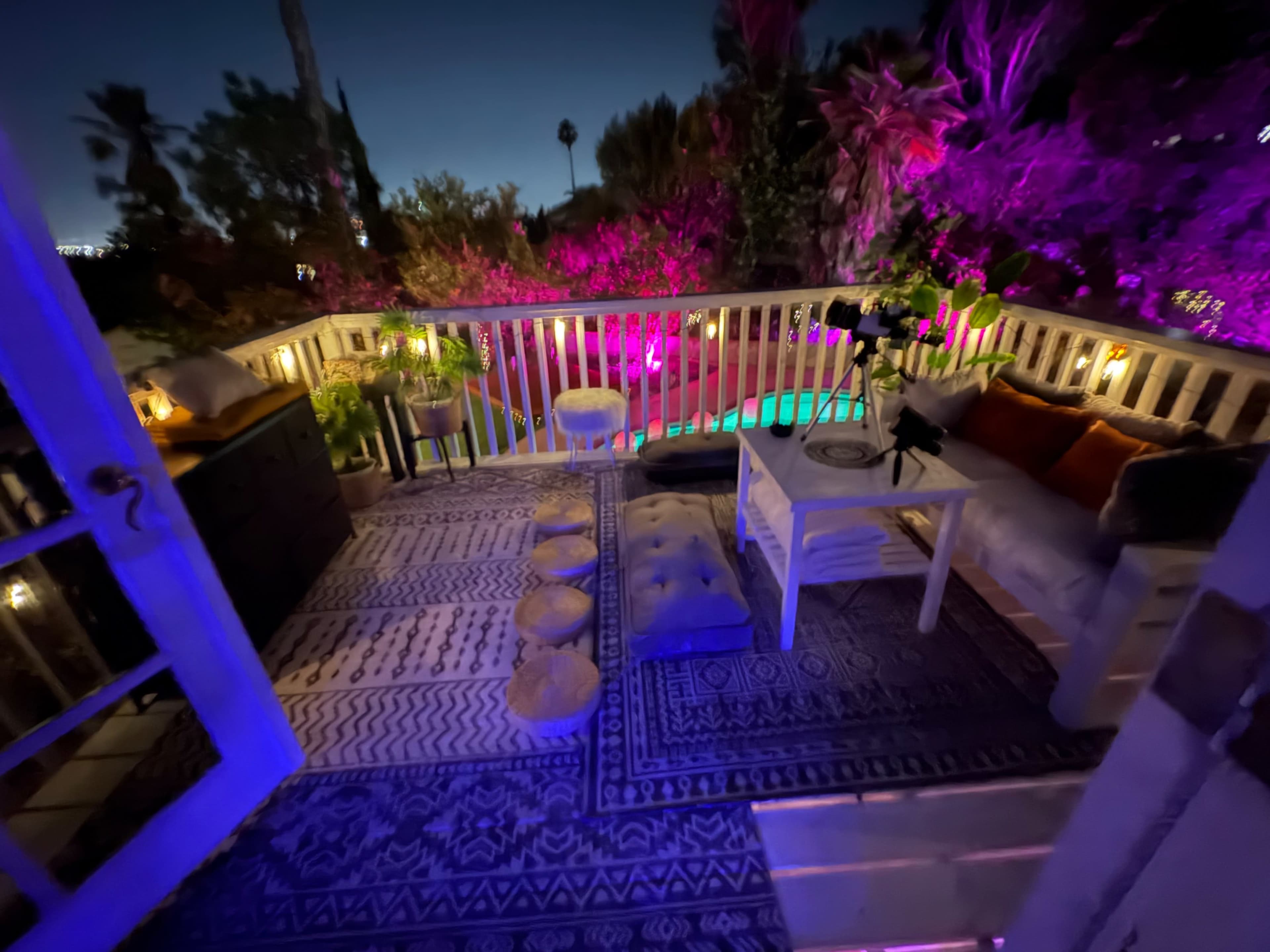 The image shows a cozy outdoor seating area illuminated by colorful lights with a view of foliage and a pool.