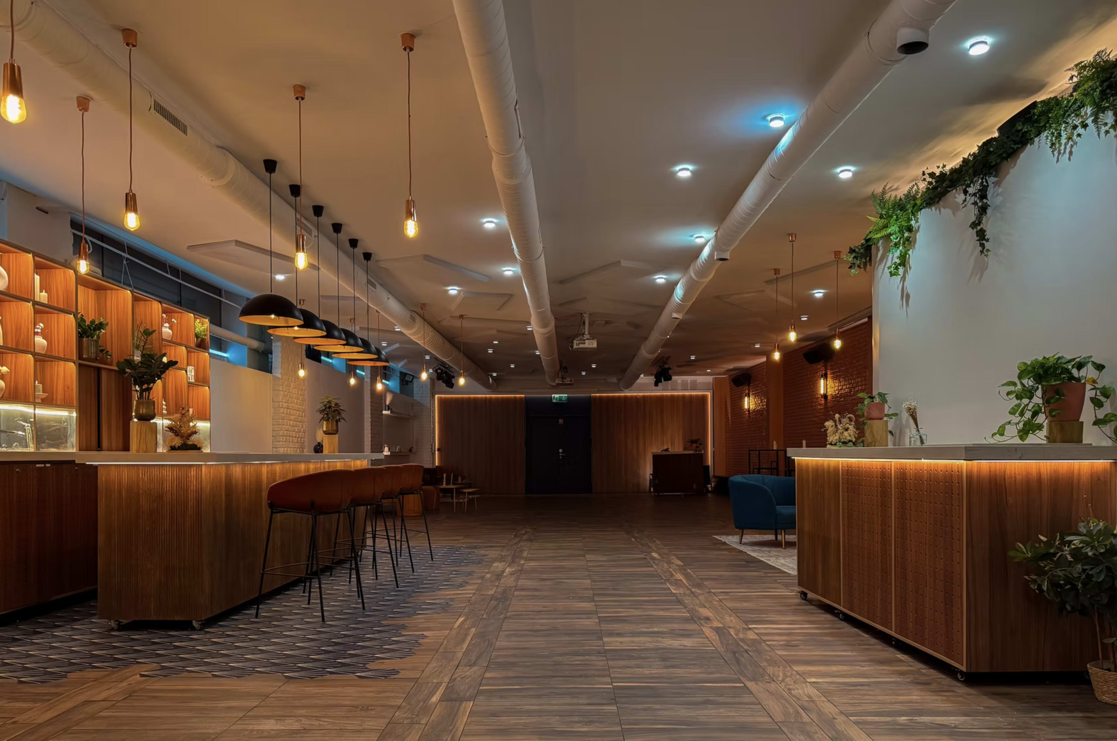The image shows a modern, well-lit interior space featuring wooden counters, pendant lighting, and a mix of seating areas with greenery and decorative plants.