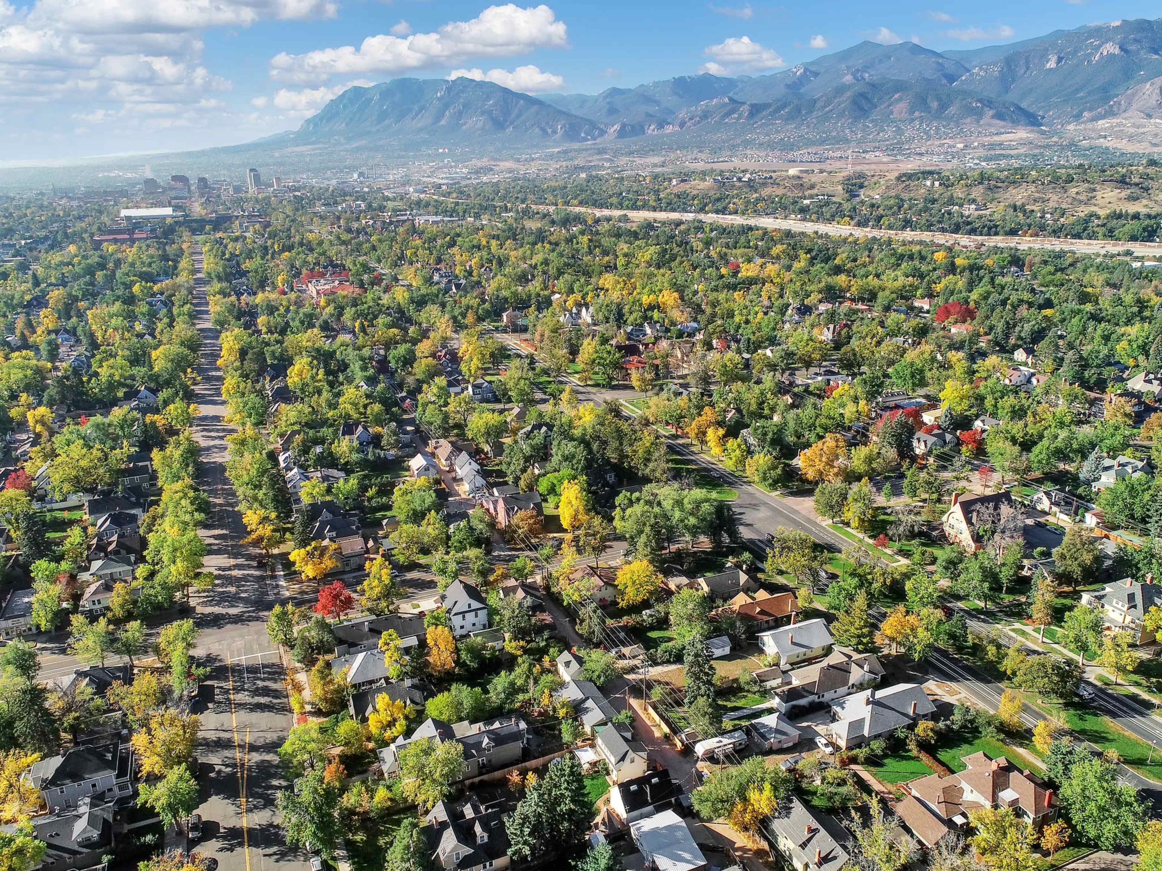 An aerial view shows a residential area with a grid layout surrounded by trees exhibiting fall colors and mountains in the background.