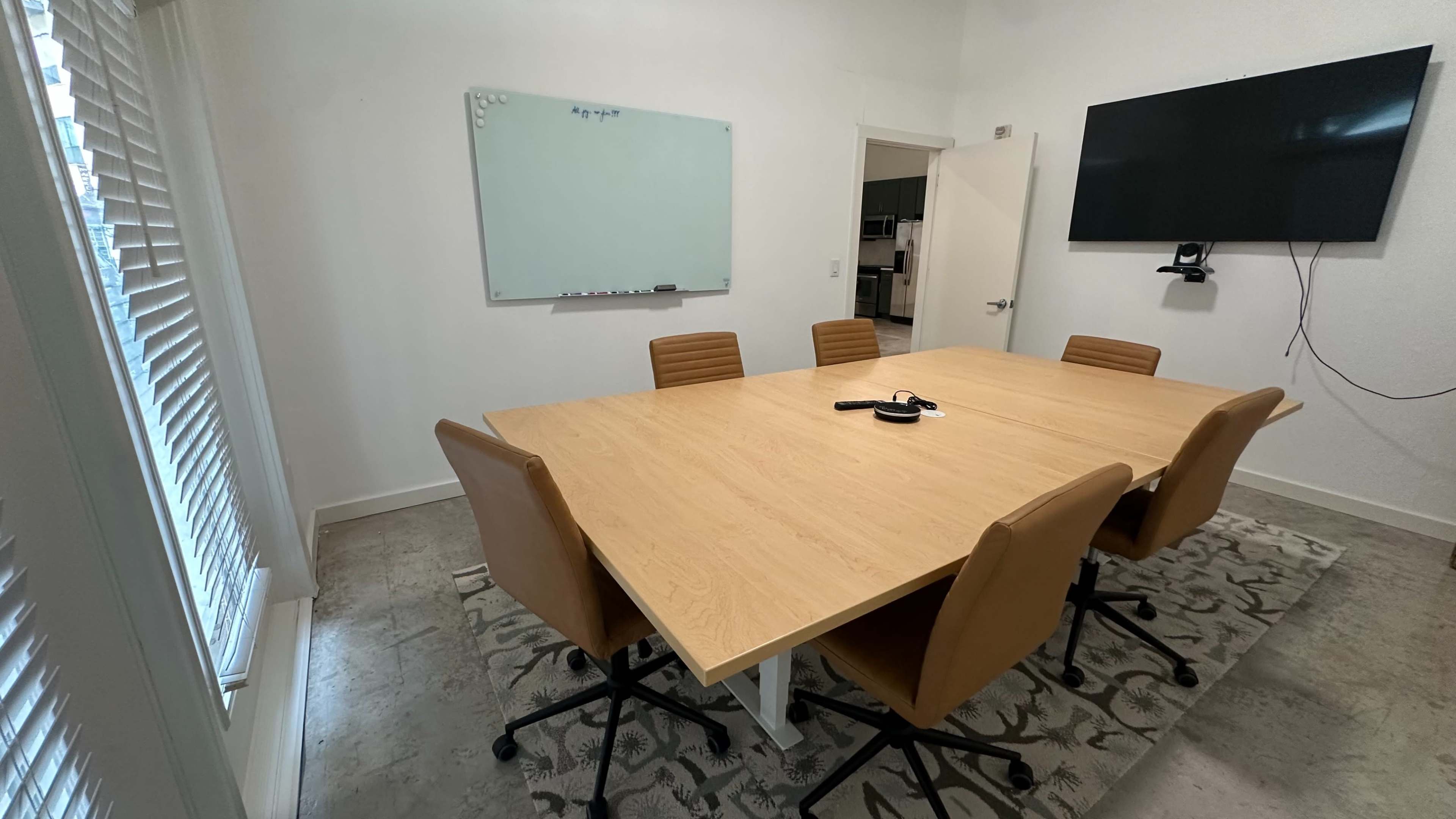 A modern conference room features a large wooden table surrounded by six brown chairs, with a whiteboard and a wall-mounted screen visible.