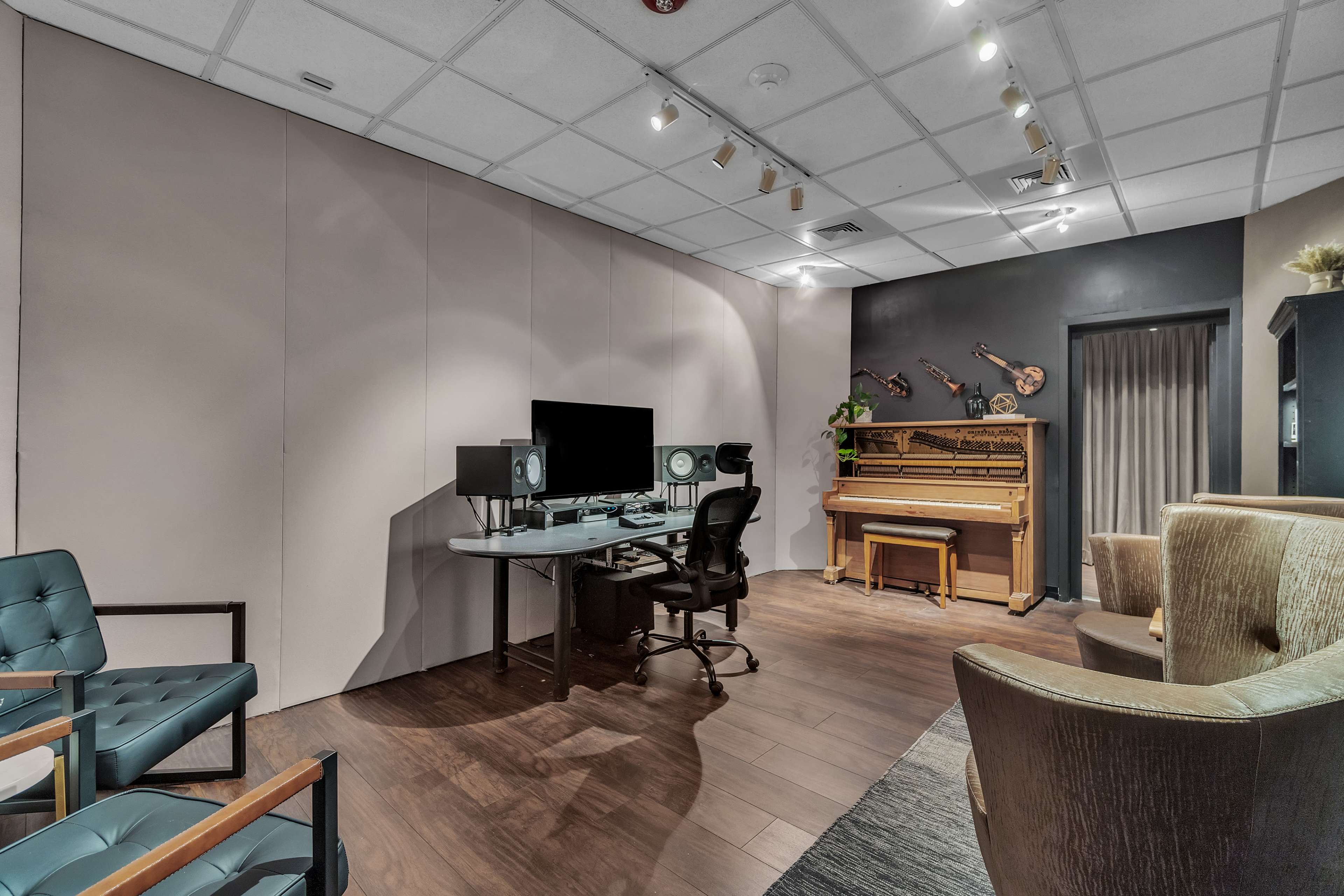 The image shows a modern recording studio with a desk equipped with audio monitors and a computer, a vintage piano in the corner, and seating arranged along the sides.